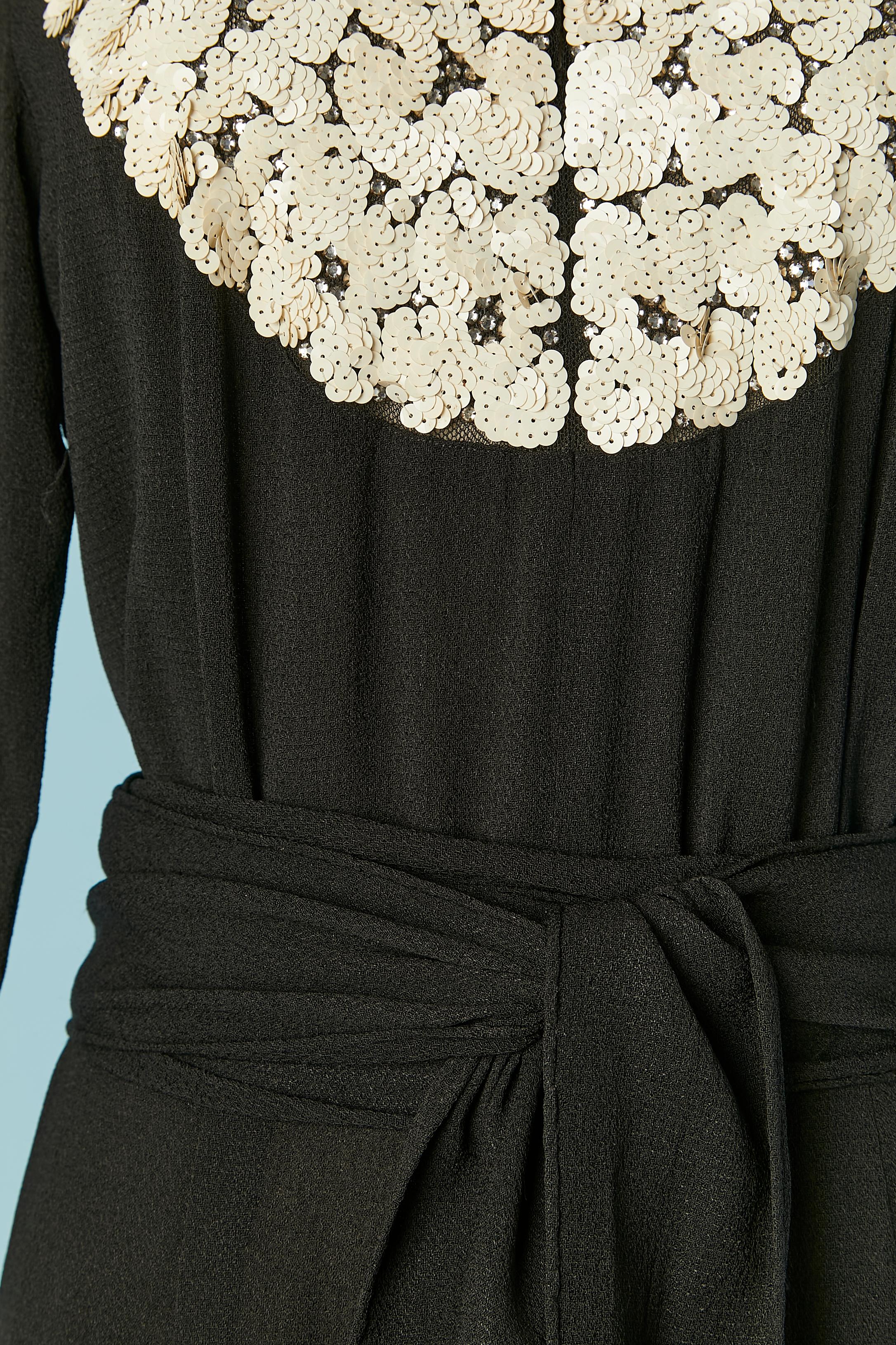 Schwarzes Abendkleid aus Crêpe mit Strass- und Paillettenverzierung auf einem durchsichtigen Tüllunterteil. Schulterpolster. Haken- und Ösenverschluss in der vorderen Mitte. Gürtel aus Stoff. Reißverschluss auf der linken Seite und an den