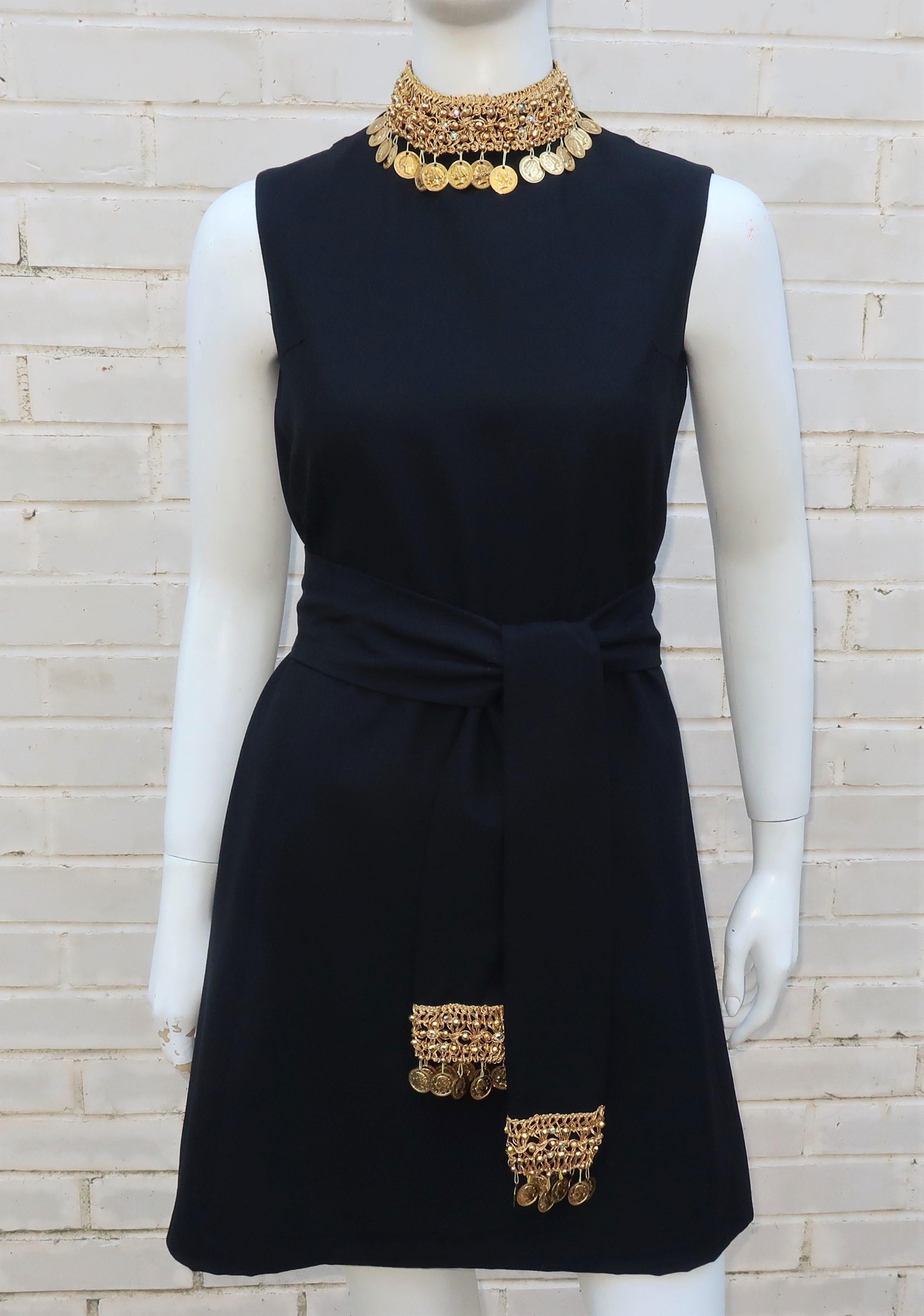 A 1960's jazzed up version of the little black dress in a black crepe fabric with gold braiding at the collar and sash embellished by beading, iridescent rhinestones and gold coins.  The simple a-line sleeveless mini dress silhouette zips and hooks