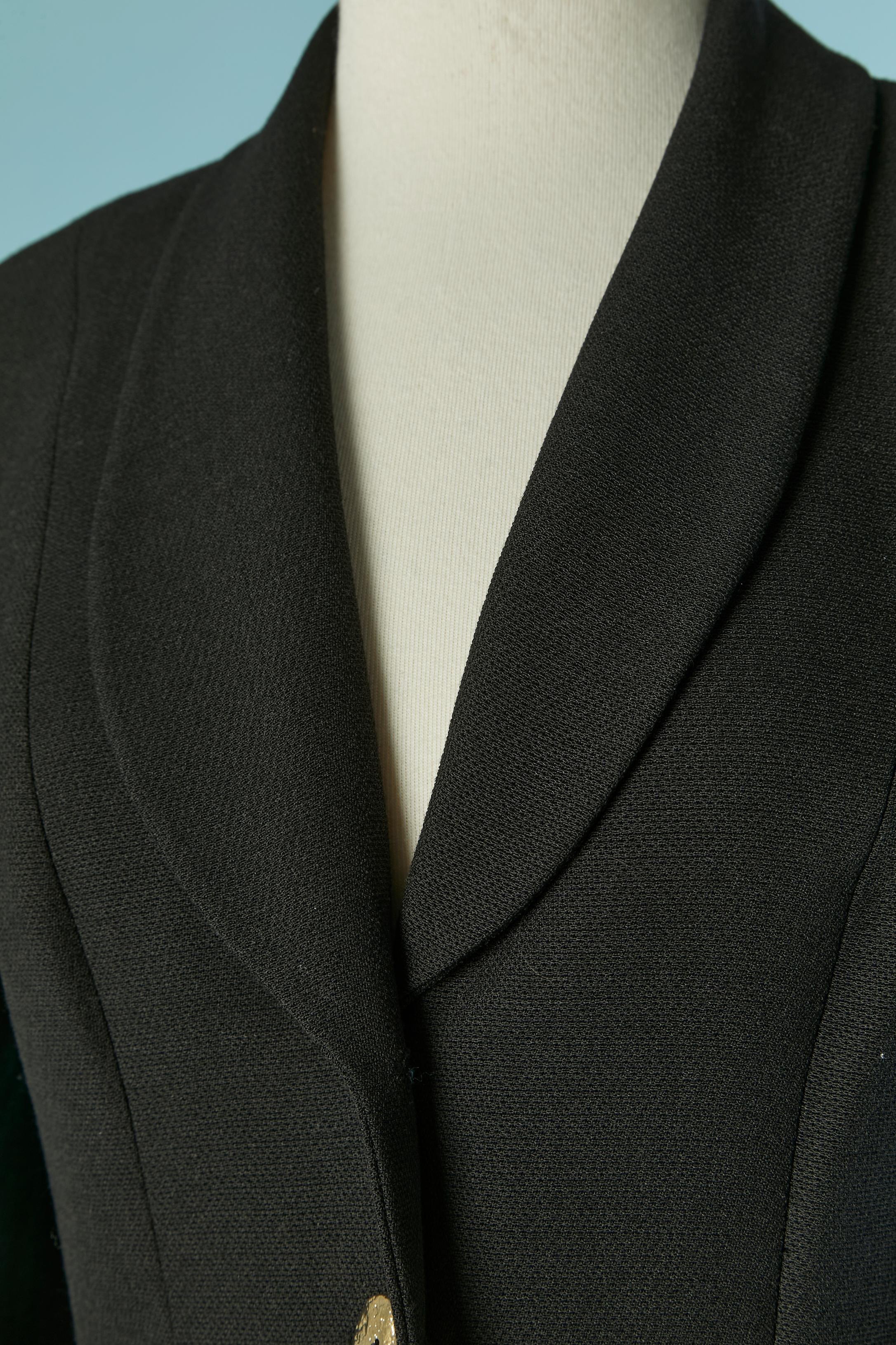 Abito con gonna in crêpe nero con spacco su giacca e gonna. Non c'è l'etichetta della composizione del tessuto, ma la fodera è probabilmente in acetato o rayon. 
Chiusura con bottoni e asole in metallo dorato sul centro della giacca. Spallaccio.