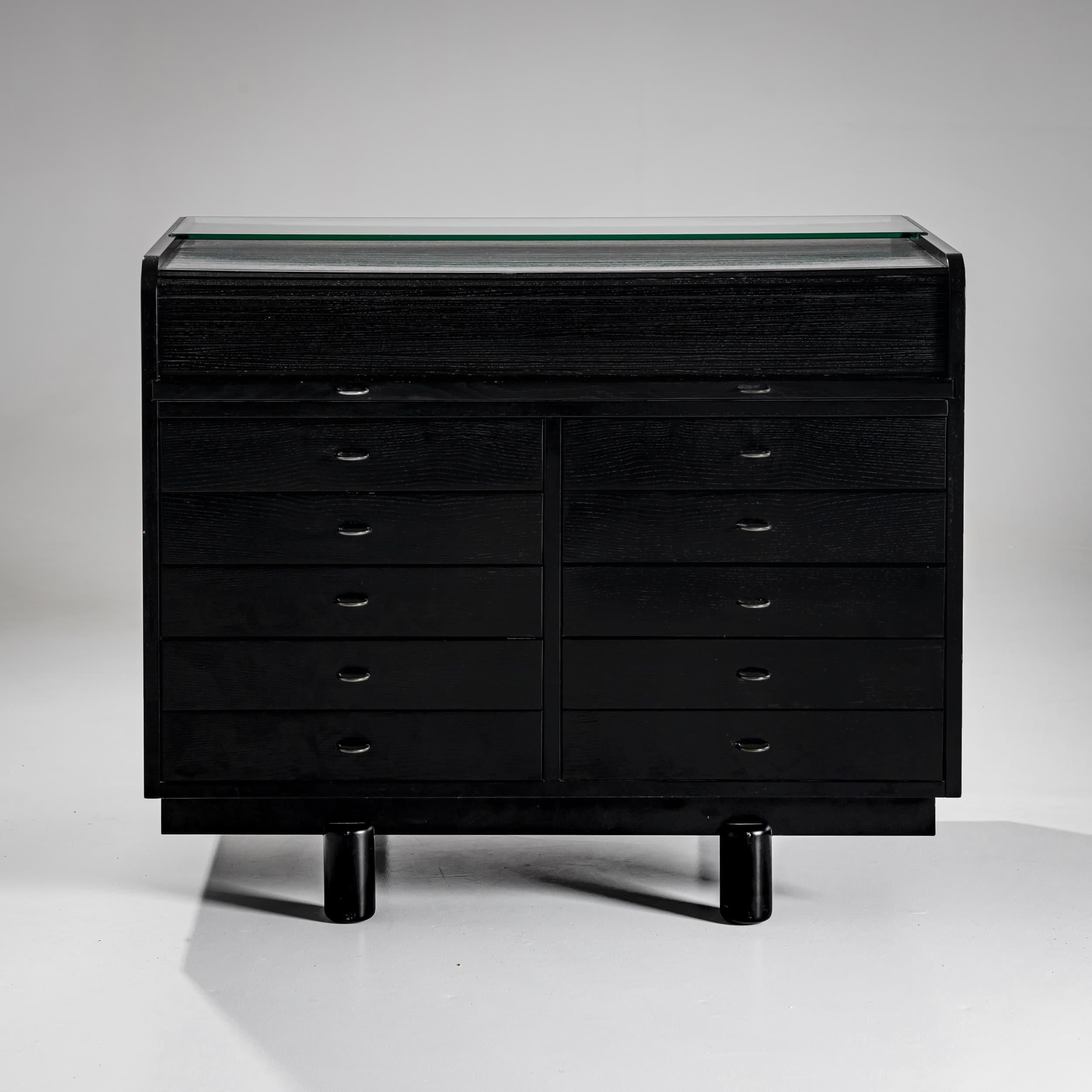 Refined black-lacquered desk on cylindrical legs, designed by Gianfranco Frattini for Bernini. The desk features ten drawers, a pull-out writing surface, and a tambour top that conceals the upper compartment.
Inside, additional small drawers are