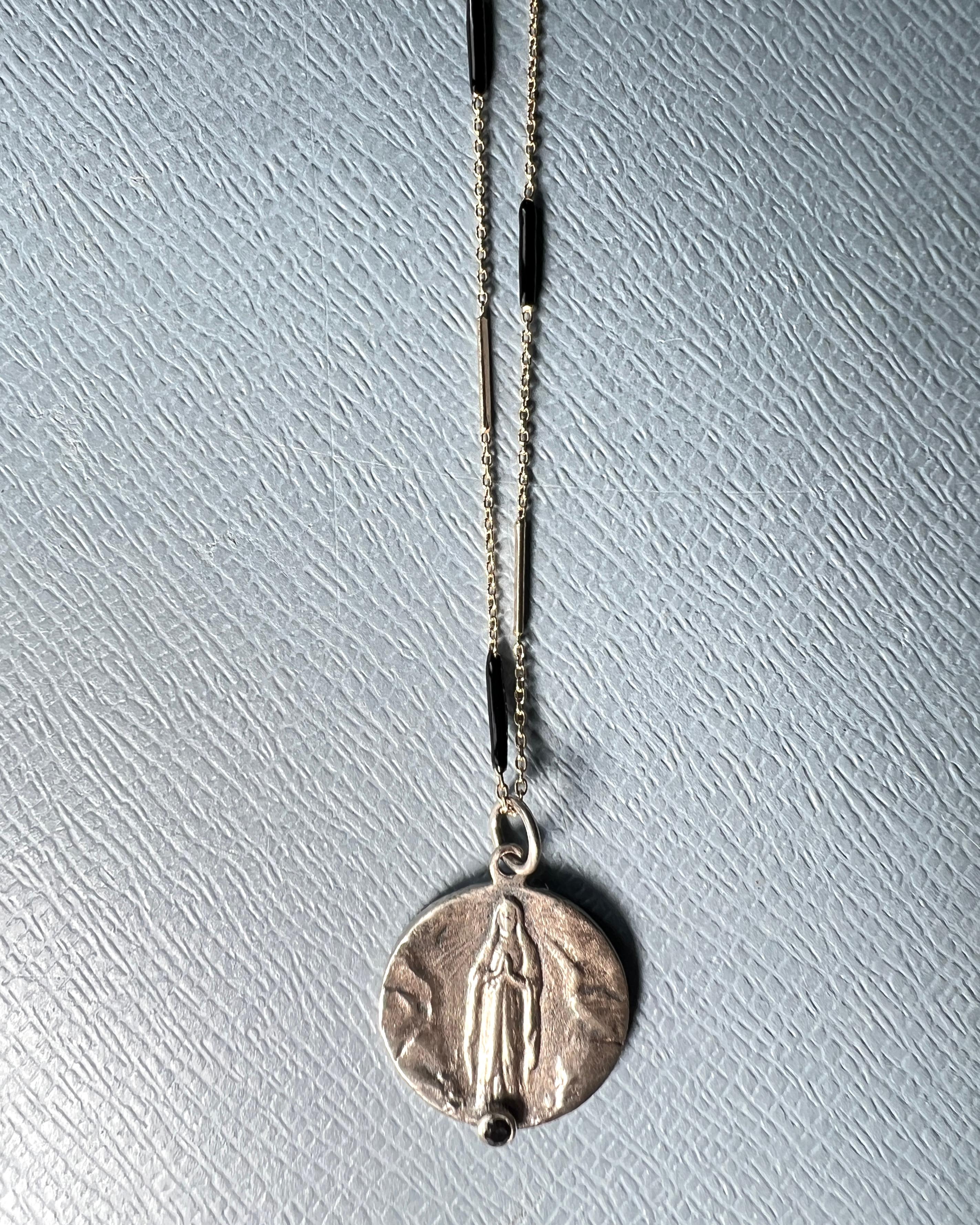 This Virgin Mary pendant necklace features a French-inspired religious coin cast in silver, accented with a natural black diamond at the base. Suspended from a black enamel chain, the piece blends devotional symbolism with modern, minimalist