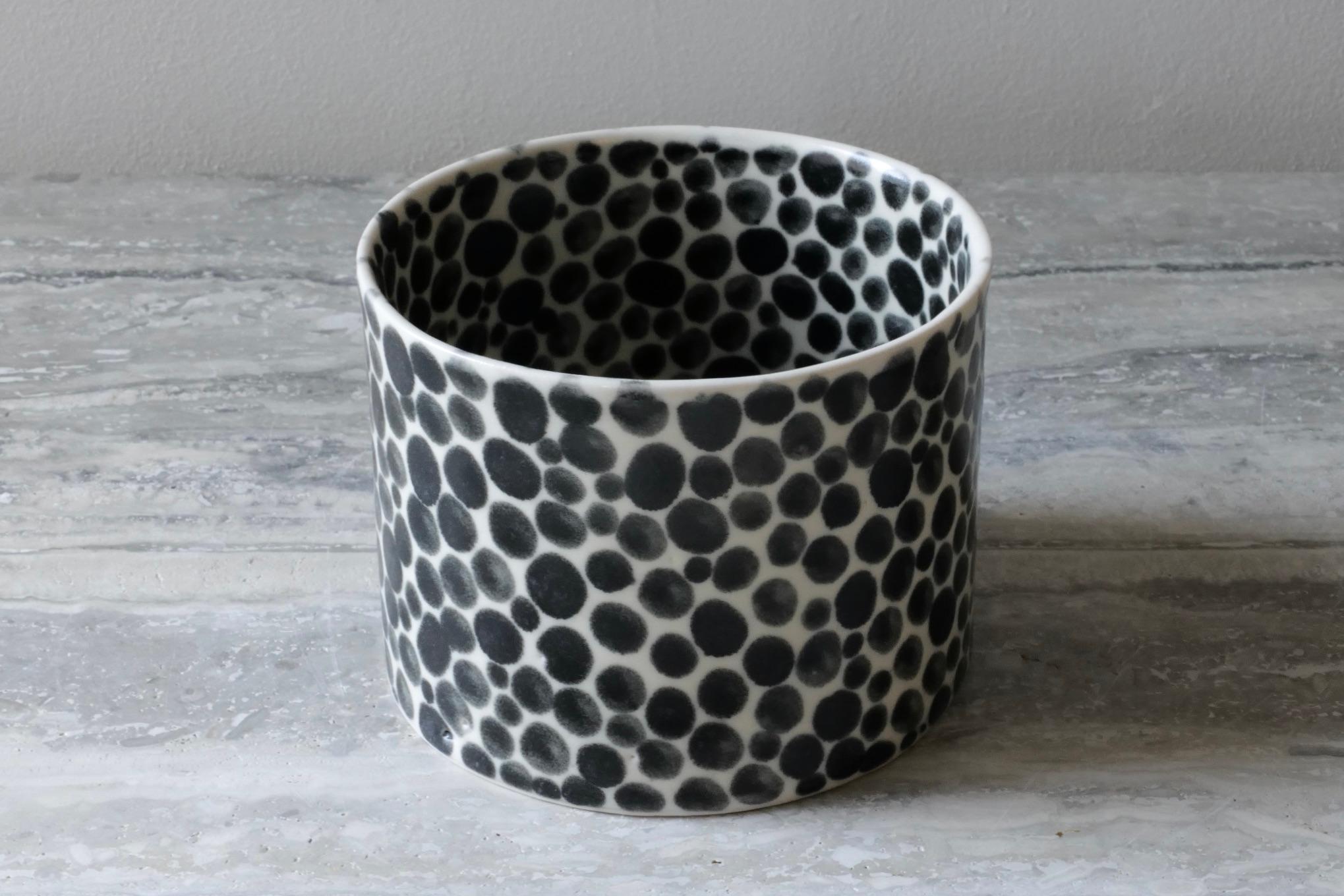 Small, short ceramic vase. Hand-cast in porcelain and once bisque fired, each dot is hand-painted with a shiny black glaze. An unconventional layered glazing technique, developed by the artist, is used in these cast porcelain pieces. The straight