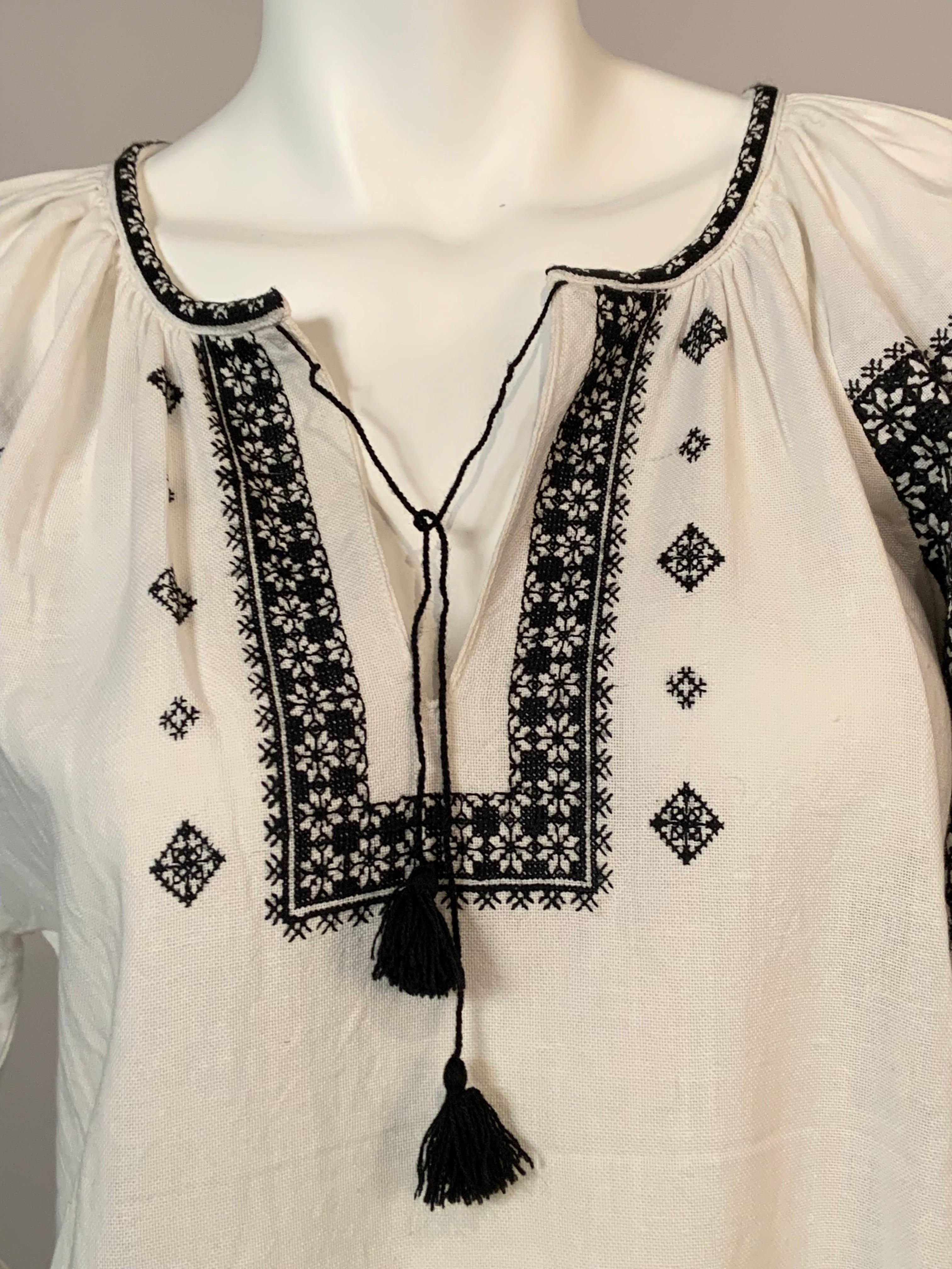 Il cotone bianco è splendidamente ricamato a mano in nero sulla scollatura, sul davanti della camicetta e sulle maniche, che sono riccamente ricamate.  La blusa si indossa sopra la testa, con un laccetto decorato con nappe sulla scollatura.  È in