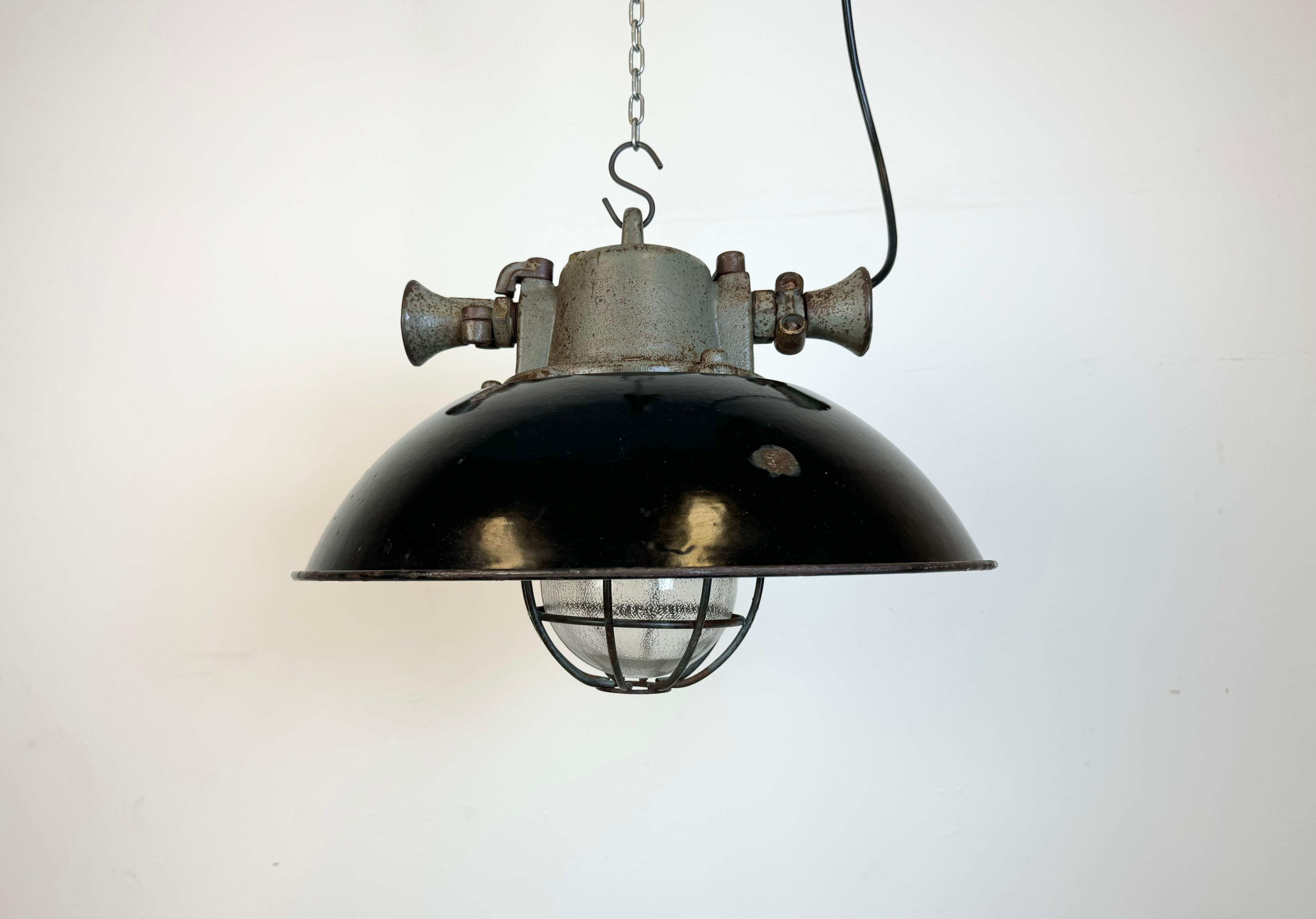 Industrial hanging lamp manufactured during the 1950s in former Czechoslovakia. It features a black enamel shade with white enamel interior, a cast iron top,a clear glass cover and an iron grid. Porcelain socket requires standard E27/E26 lightbulbs.