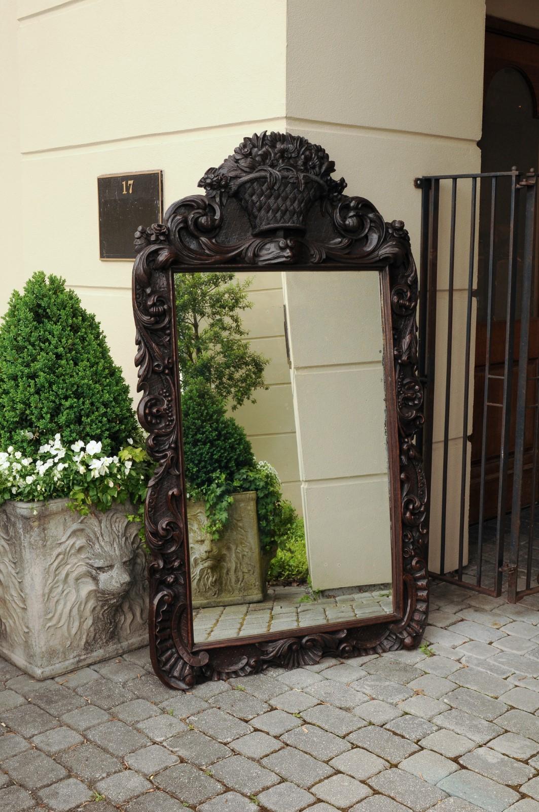 A hand carved wooden mirror from the early 20th century, with dark patina, floral decor and scrolls. Born in the early years of the 20th century, this exquisite Black Forest mirror features a sinuous Silhouette, accented with delicate scrolls