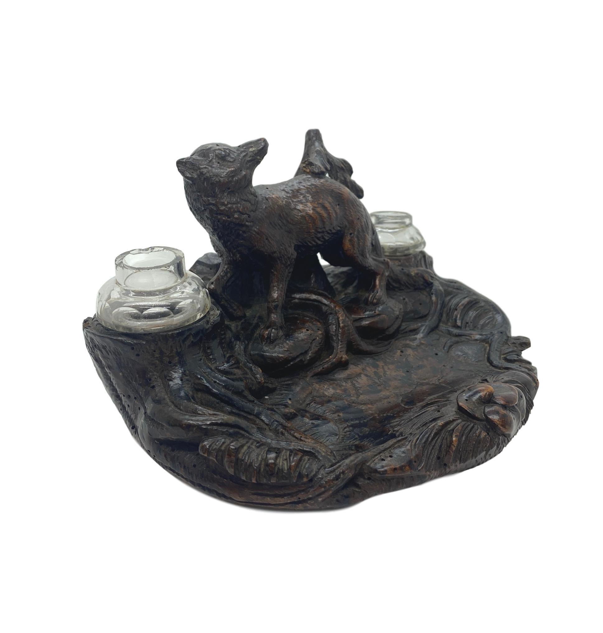 Black Forest Carved Inkstand, naturalistically hand-carved of solid linden wood, with the central figure of a fox among a rootwork base simulating a forest floor, with two tree stumps forming the inkpot holders, with two glass inkwells (chips to