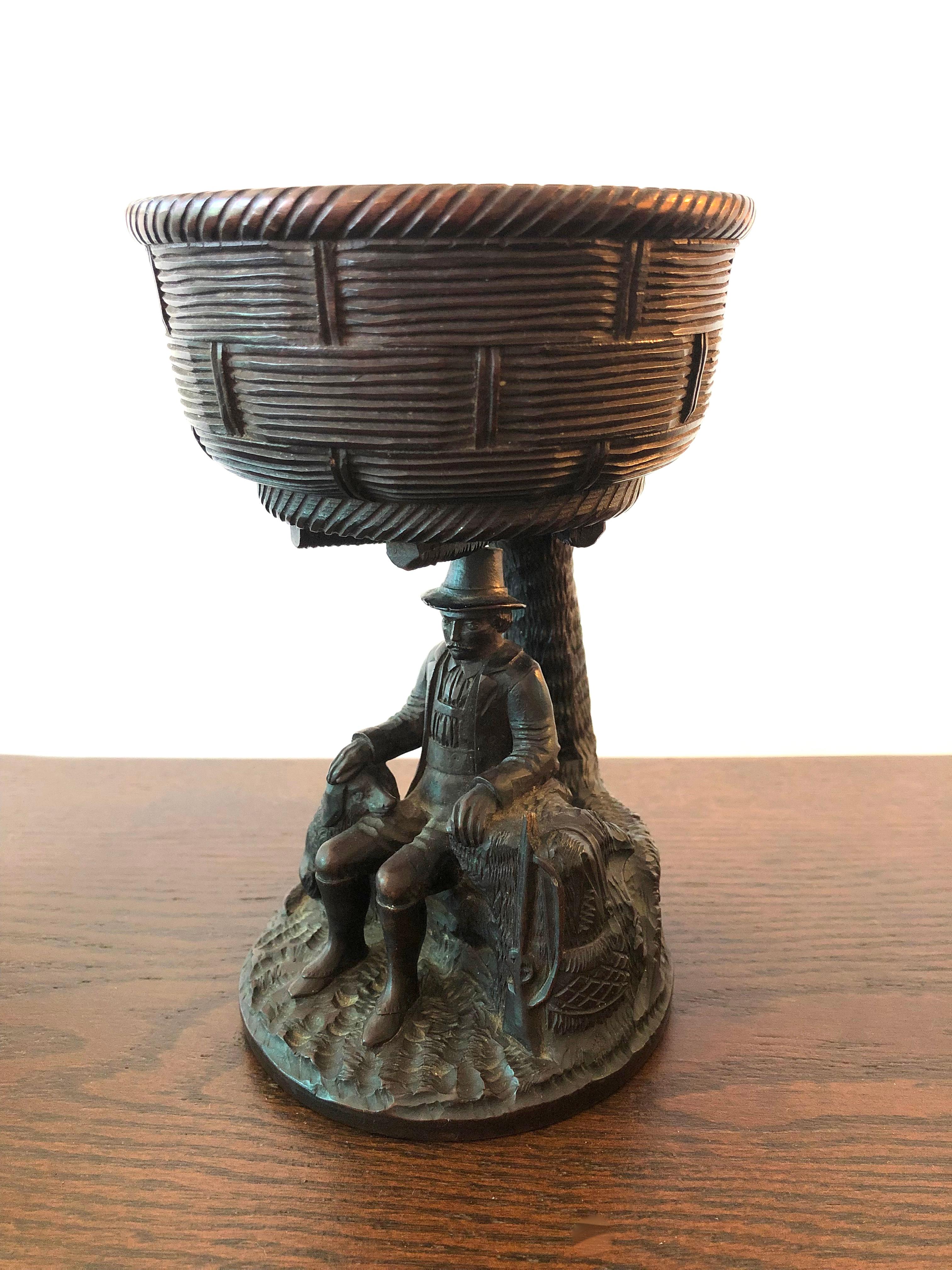 A beautifully crafted Black Forest hand carved walnut hunting presentation cup, the basket weave bowl supported by the figure of a gentleman hunter leaning against a tree trunk with his gun at one side and his hand on the head of his faithful dog