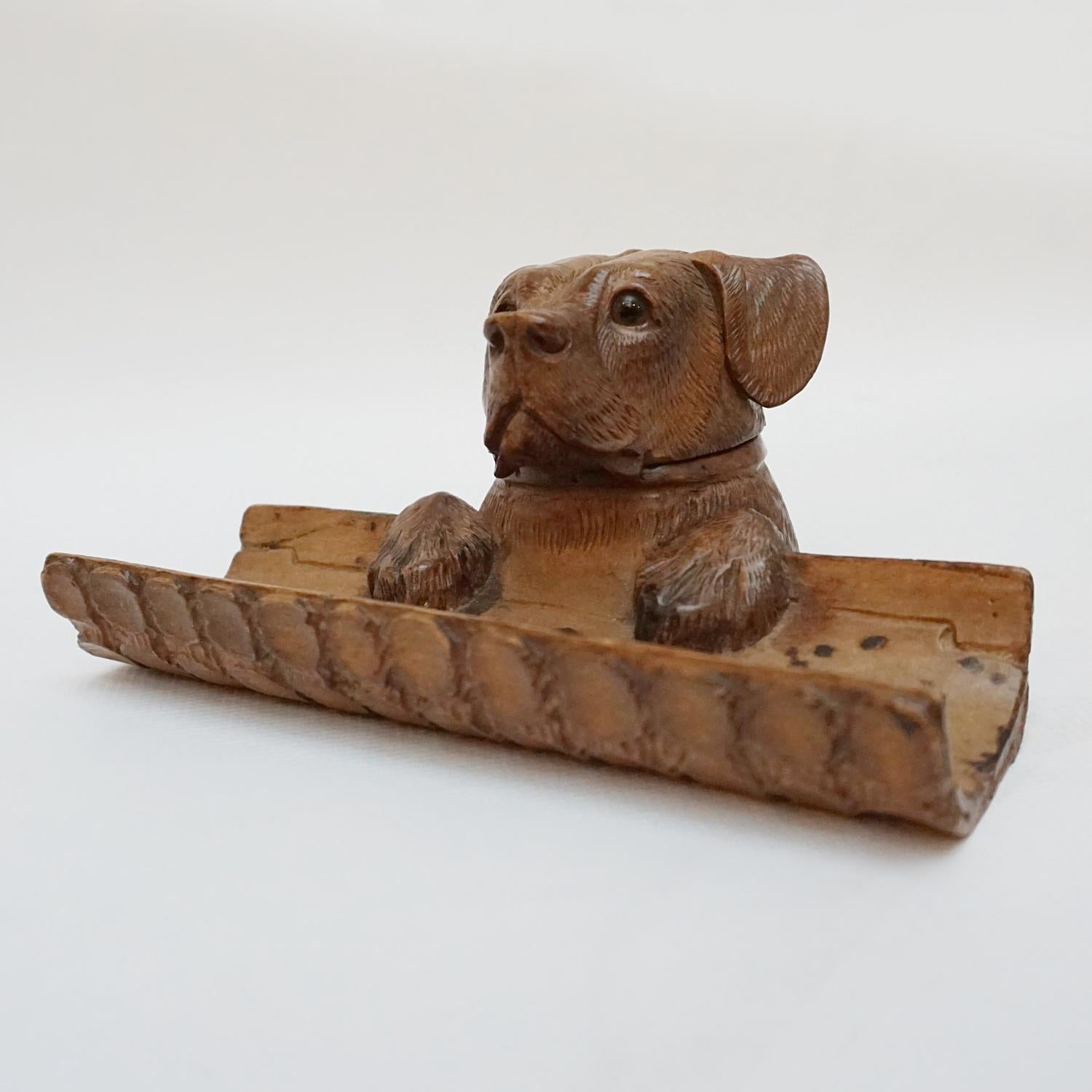 Black Forest dog inkwell and pen holder. A young dog with his paws on a piece of bark peering over the edge. Carved Linden wood. The head of the dog opens to reveal inkwell. 

Dimensions: H 9cm W 19cm D 12cm 

Origin: Swiss

Date: Circa