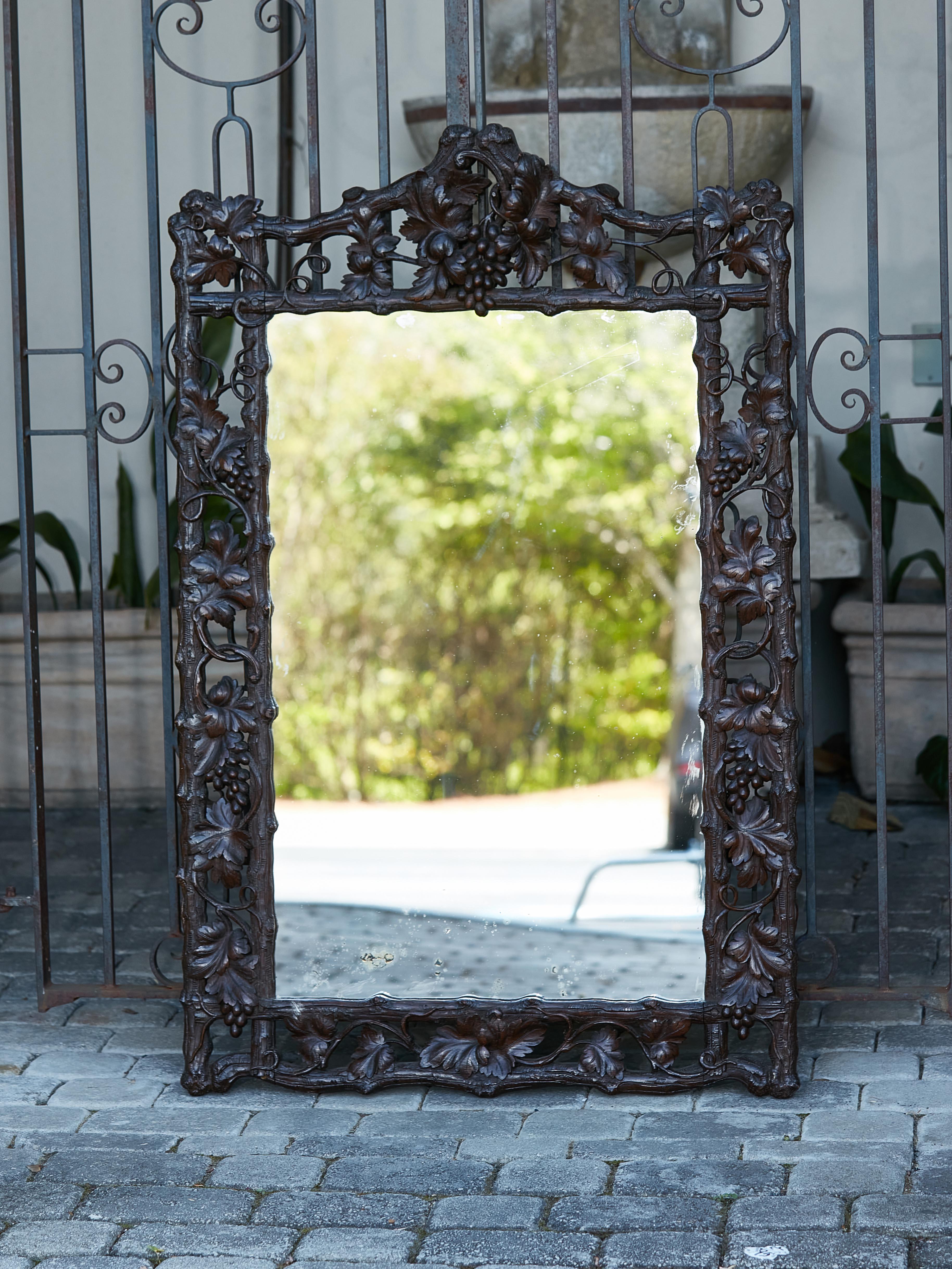 A Black Forest mirror from the Turn of the Century with hand-carved grapevine motifs and dark patina, circa 1900. Emanating a symphony of rustic charm and skilled artistry, this Turn of the Century Black Forest mirror, circa 1900, is an emblem of