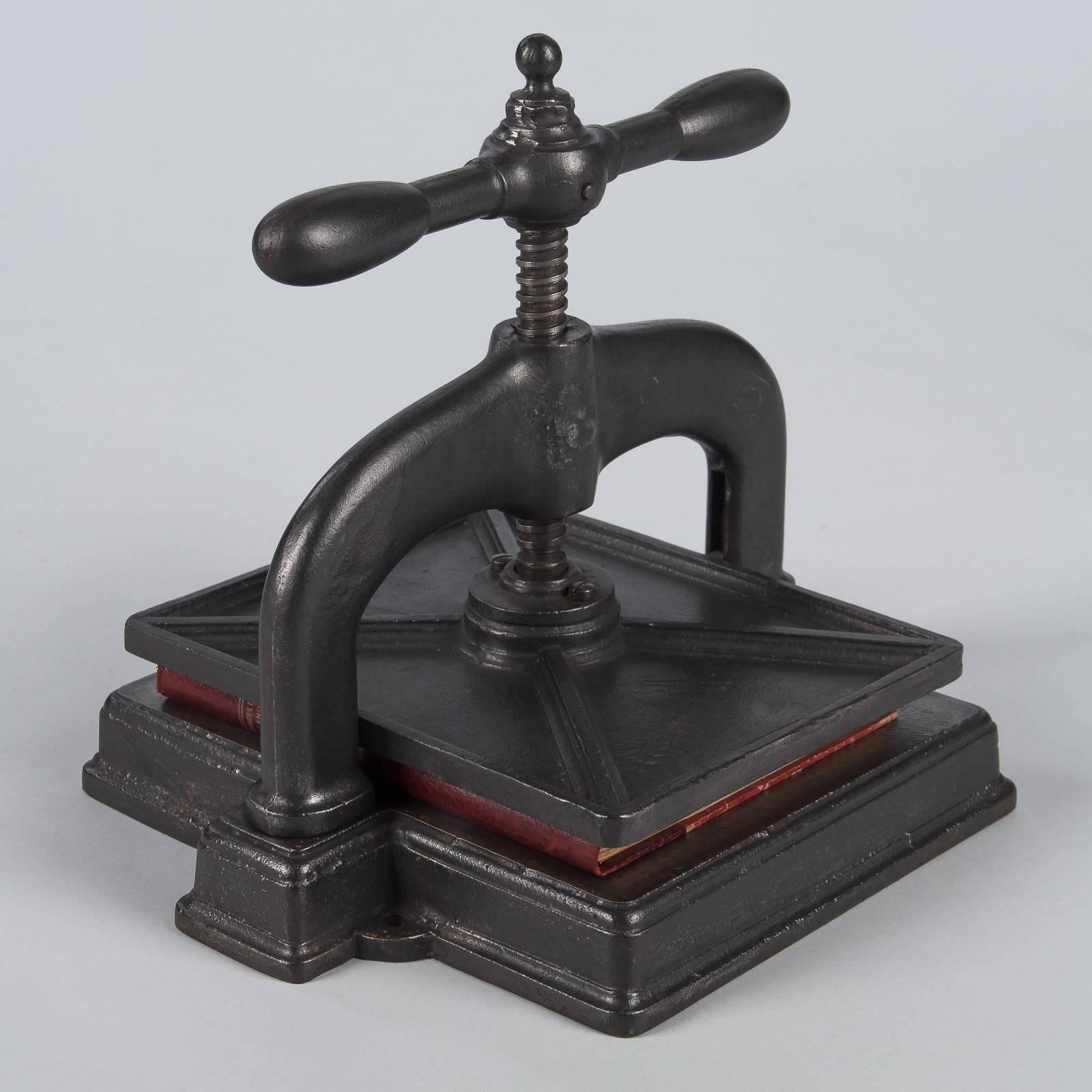 A black forged iron Industrial book press from the early 1900s. Beautiful patina on the iron. Squat shape with an Industrial aesthetic. The base is square with light molding. Curved, stout shoulders house a screw mechanism with baton shaped handles