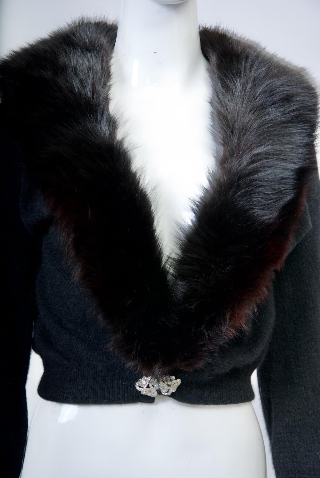 Schwarze Strickjacke aus einer Wollmischung mit abnehmbarem Fuchskragen. Die Taille ist mit einer Strassspange und die Handgelenke mit zwei Strasssteinen verziert. Der Pelzkragen lässt sich mit kleinen Druckknöpfen am Rand an- und abnehmen, wenn der