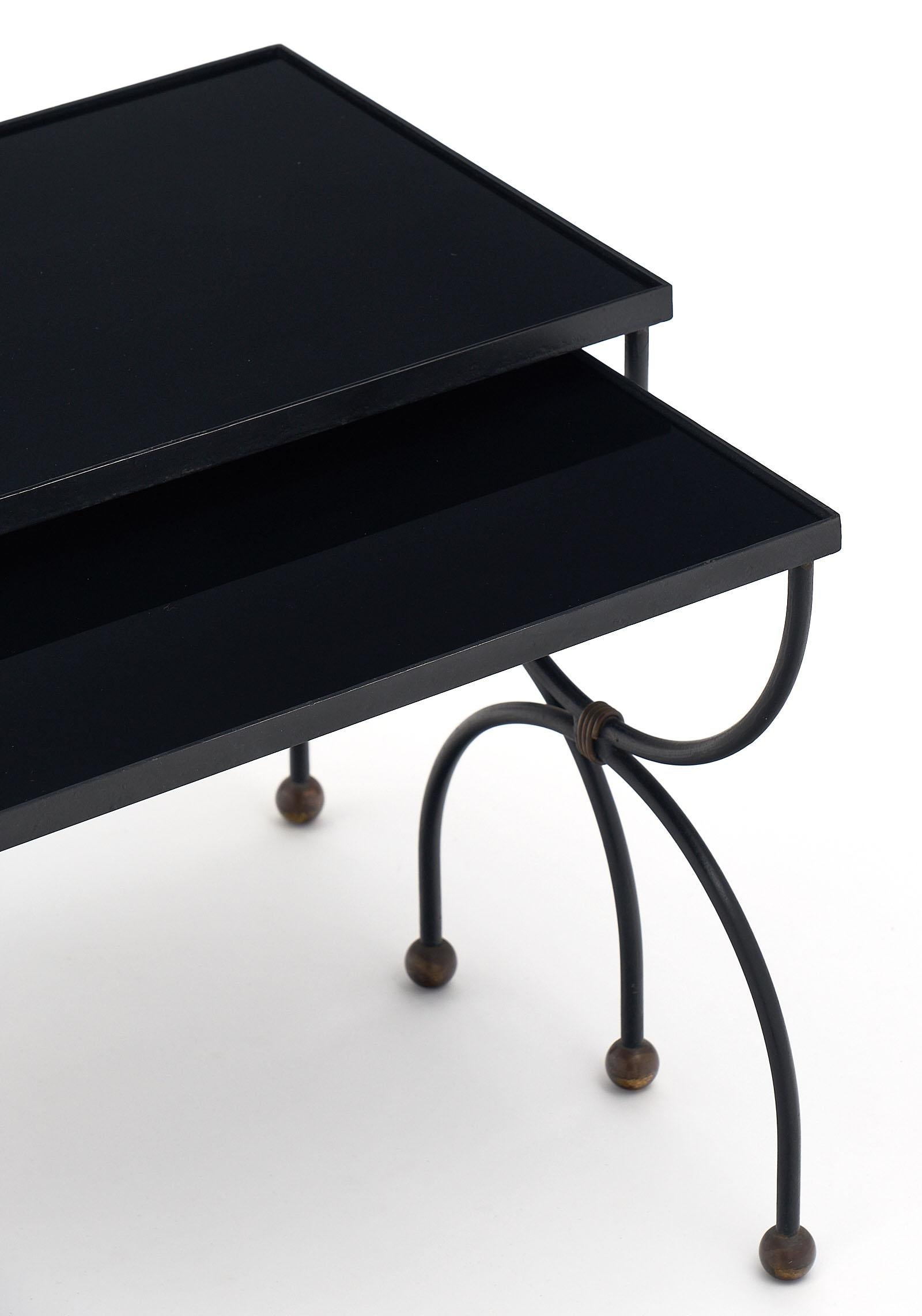 Art Deco period black glass nesting tables with steel bases featuring a curule shape and gold leaf accents. The tops are black cobalt glass. The measurements listed are for the larger table. The smaller table is 15” in height, 11.875” in depth, and