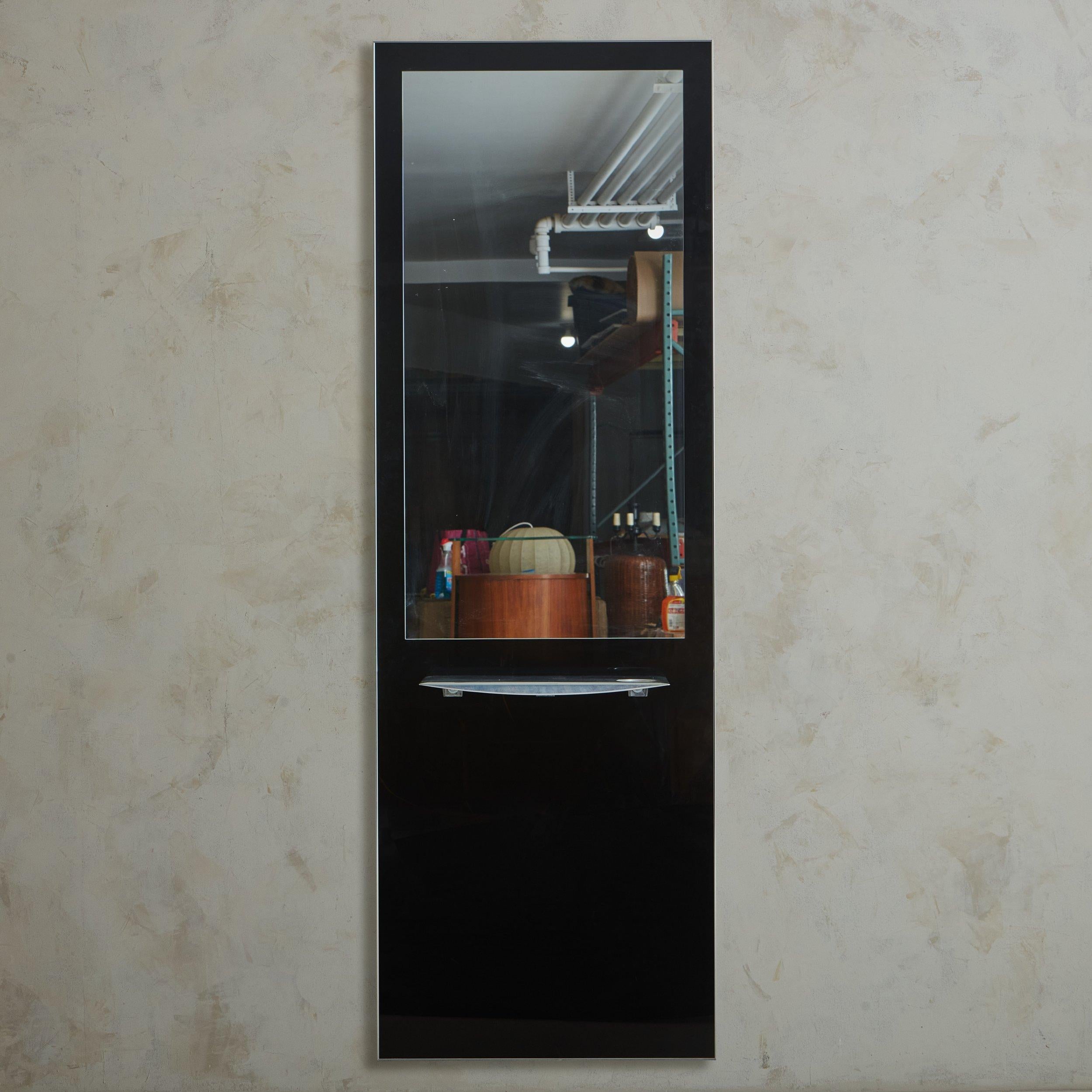 A sleek vintage French mirror featuring a dramatic wall-mount rectangular black glass frame with an inset mirror. This piece has a removable aluminum shelf with one circular storage cutout detail. We can picture it in a chic walk-in closet, powder