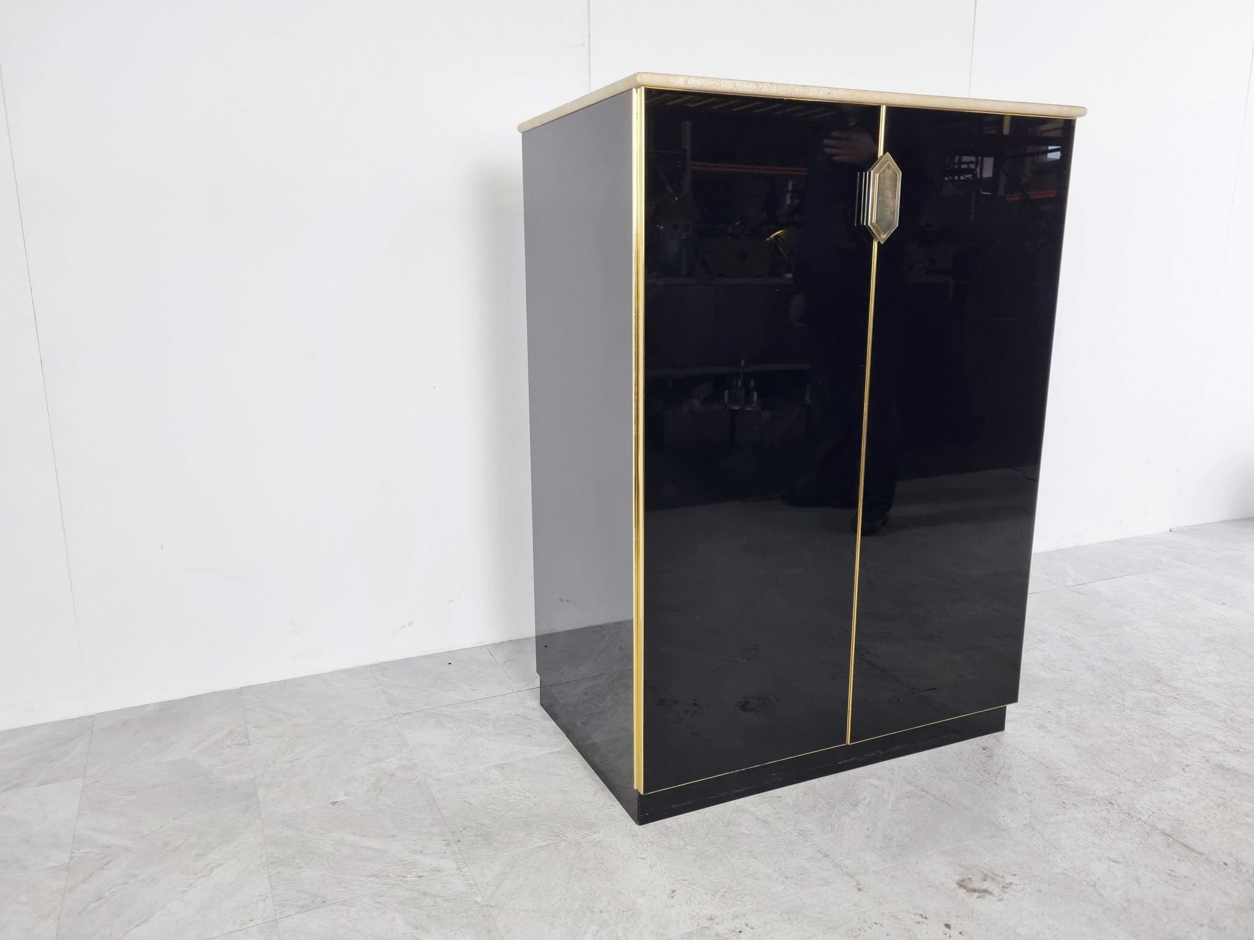 Hollywood regency black glass and brass bar cabinet with a beautiful travertine stone top.

It features 2 doors with a nicely shaped door handle and two shelves - Plenty of storage space for glasses and bottles. 

This pieces gives a great touch