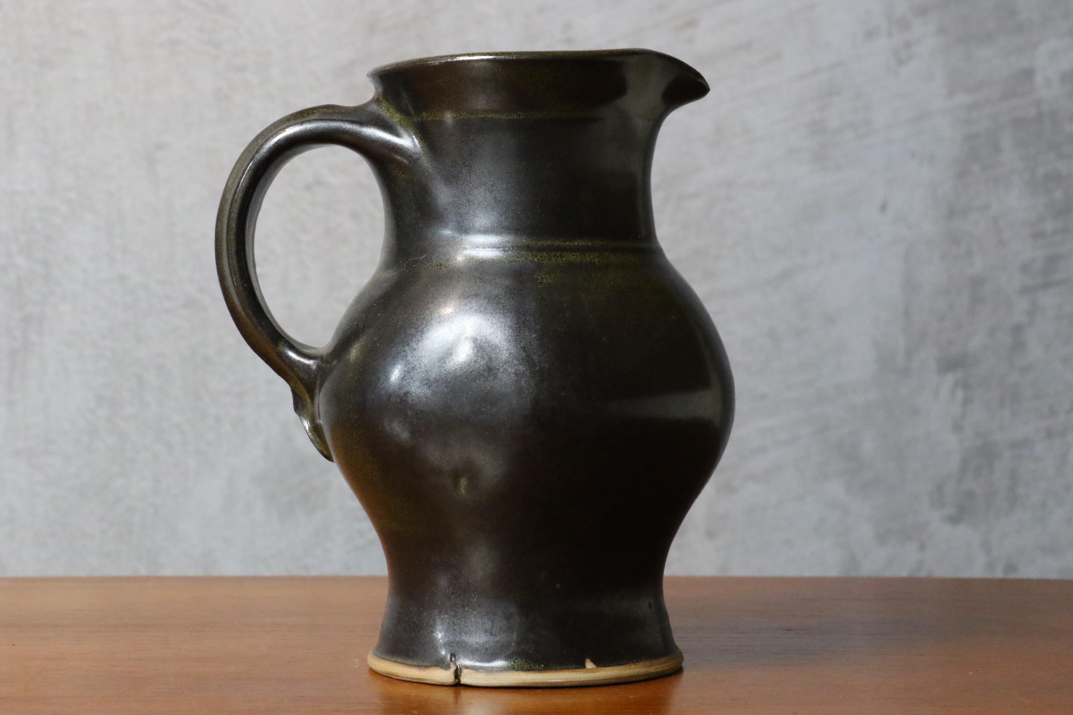 Black glazed ceramic pitcher with green glints by Jean Girel, French ceramist

This is a very beautiful ceramic pitcher. The shape of the jug and the handle remind us of the shapes of popular pottery works, while the very refined work of the
