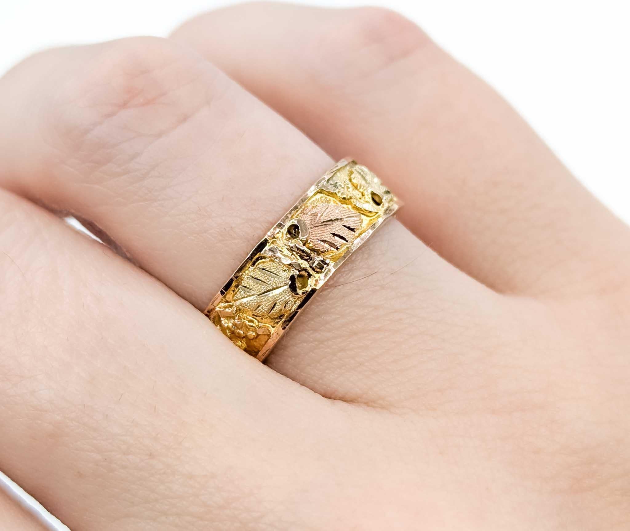 Celebrate the artistry of Black Hills Gold with this beautifully detailed tri-tone band. Crafted in 10k gold, the ring features the traditional leaf and vine motif in yellow, rose, and green gold, accented with textured details that highlight the