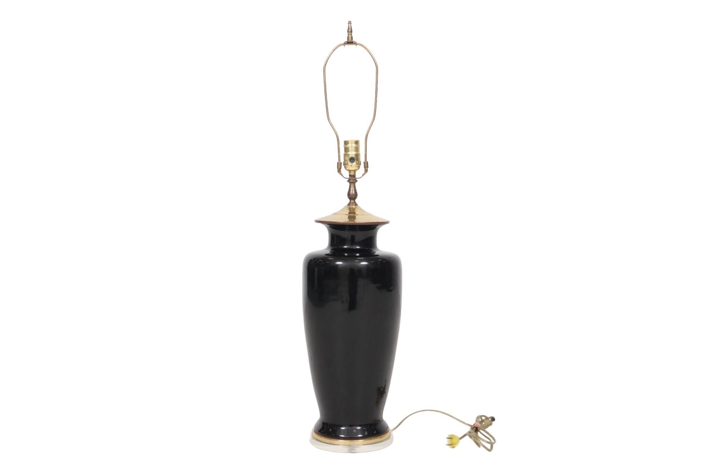 A Hollywood Regency style table lamp. The black baluster vase is ceramic with a brass cap and foot over a round lucite base. Measures 7.5”W x 7.5”D x 23.25