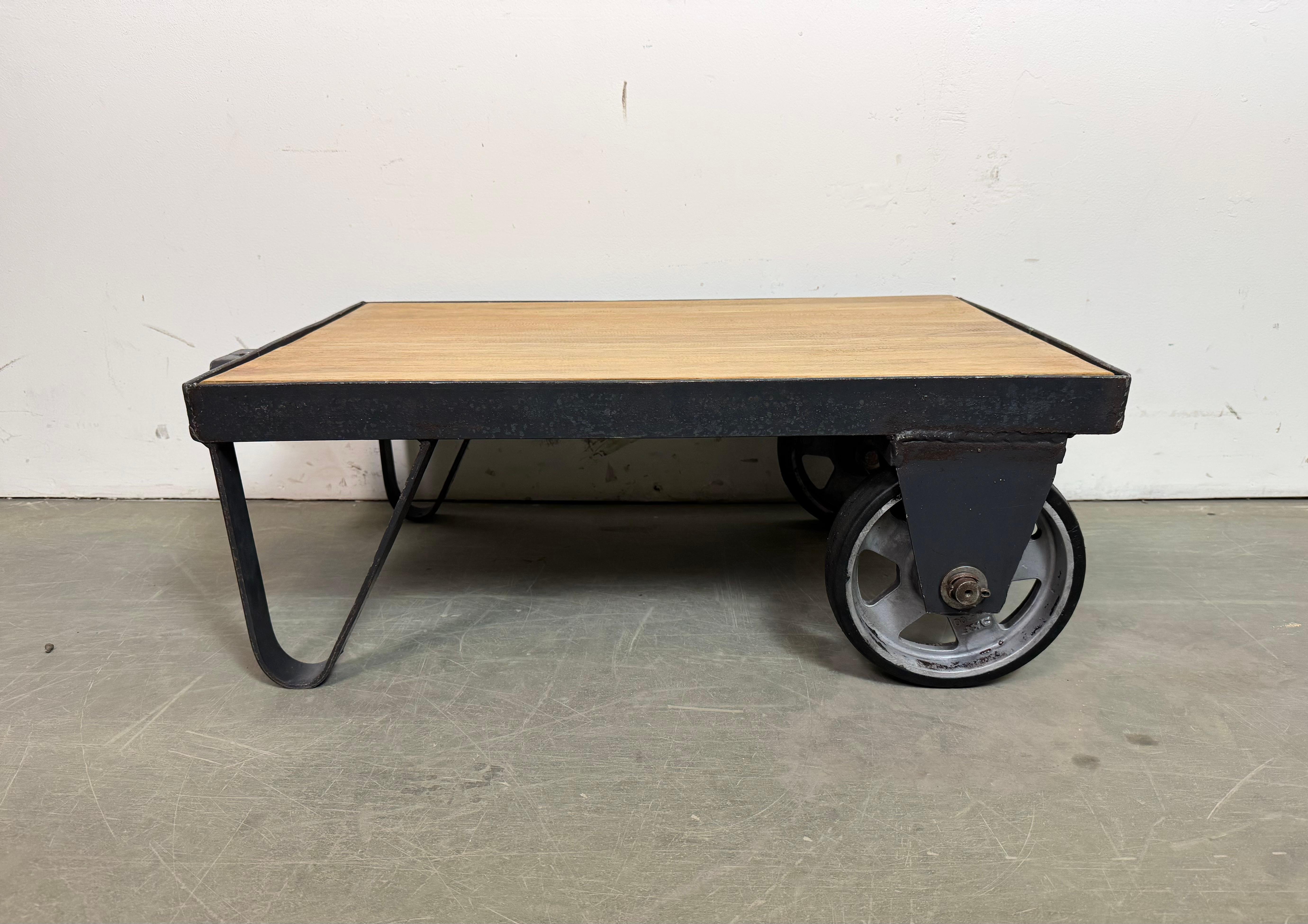 Former pallet truck from a factory now serves as a coffee table. It features a black iron construction with two original wheels and a solid wooden plate. The weight of the table is 26 kg.
