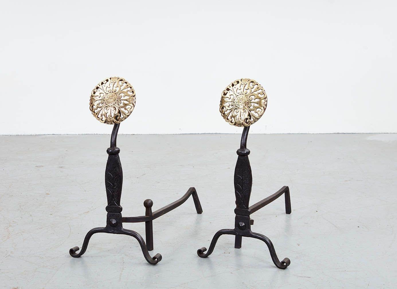 A pair of black iron andirons with sculpted and stamped shafts on arched feet ending in scrolled toes, with robustly peaned back supports, surmounted by brass rosette disks with tulip motifs applied to circular steel backplates.