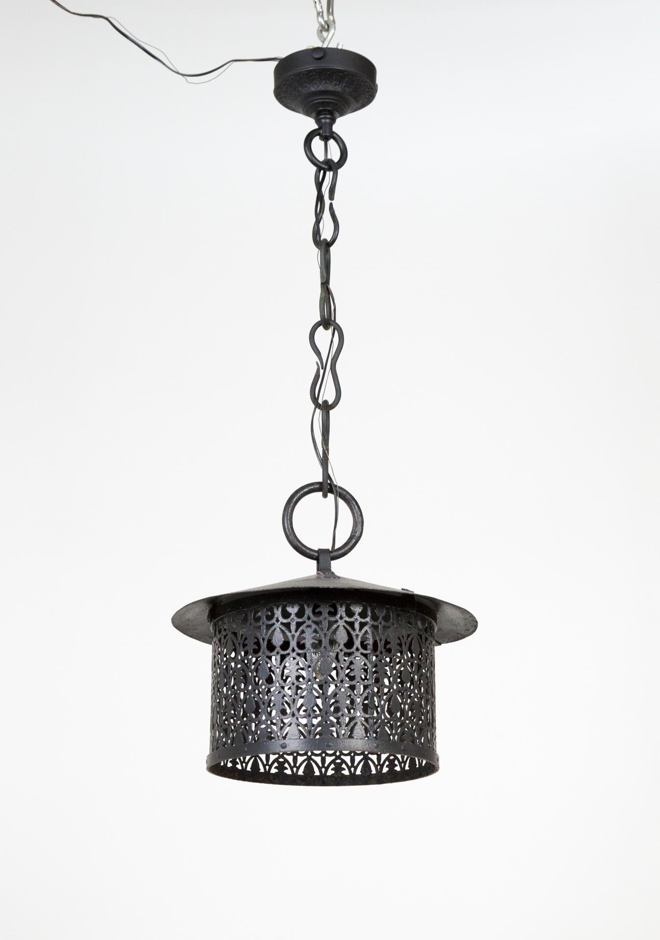 An Arts & Crafts, cylindrical, wrought iron lantern with decorative cut shapes, stylized acorn and tree details; painted black. Asian influenced in its simplicity. A large loop accent on a unique, hand forged chain with a decorative canopy.