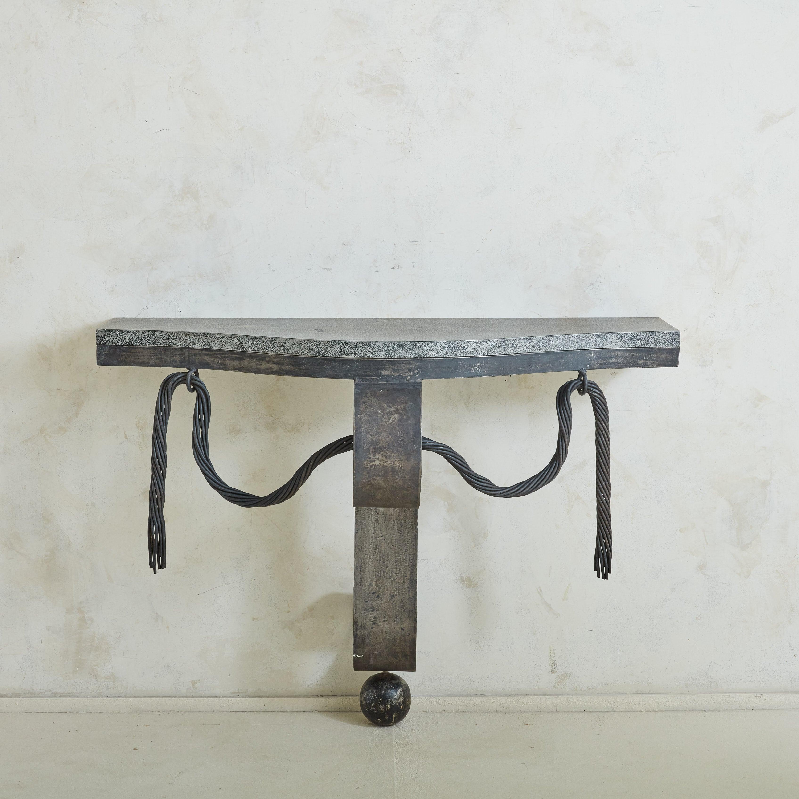A 1980s Art Deco wall-mounted console table featuring a patinated black iron base and demilune wood veneer top. The elegant triangular frame is composed of a large scroll support, decorative draped rope motifs, and an oversized ball detail along the