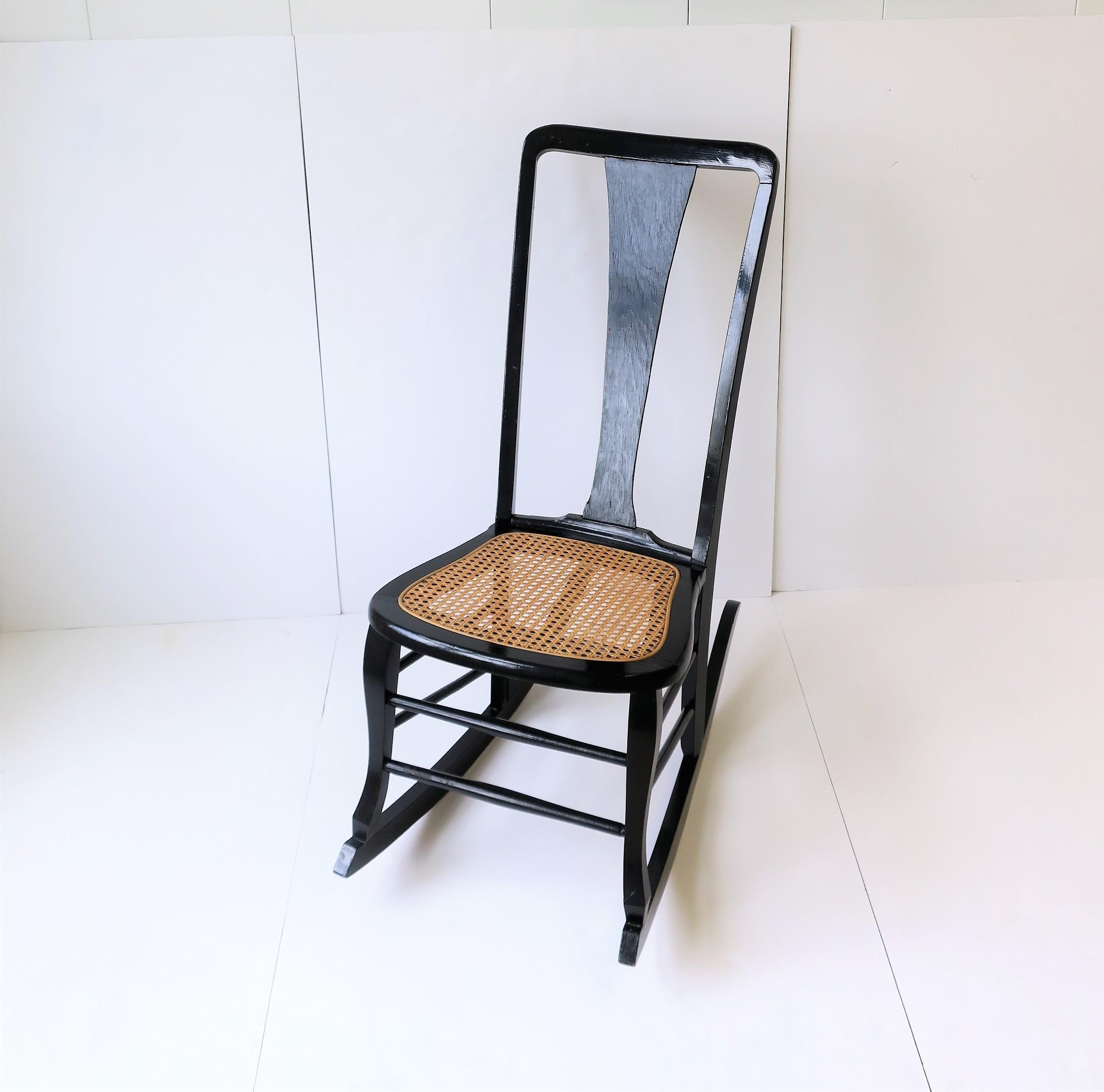 A black painted wood and blonde cane rocking chair, circa mid-20th century. Wood chair is a painted black lacquered style with a blonde cane seat. Chair measures: 28.5