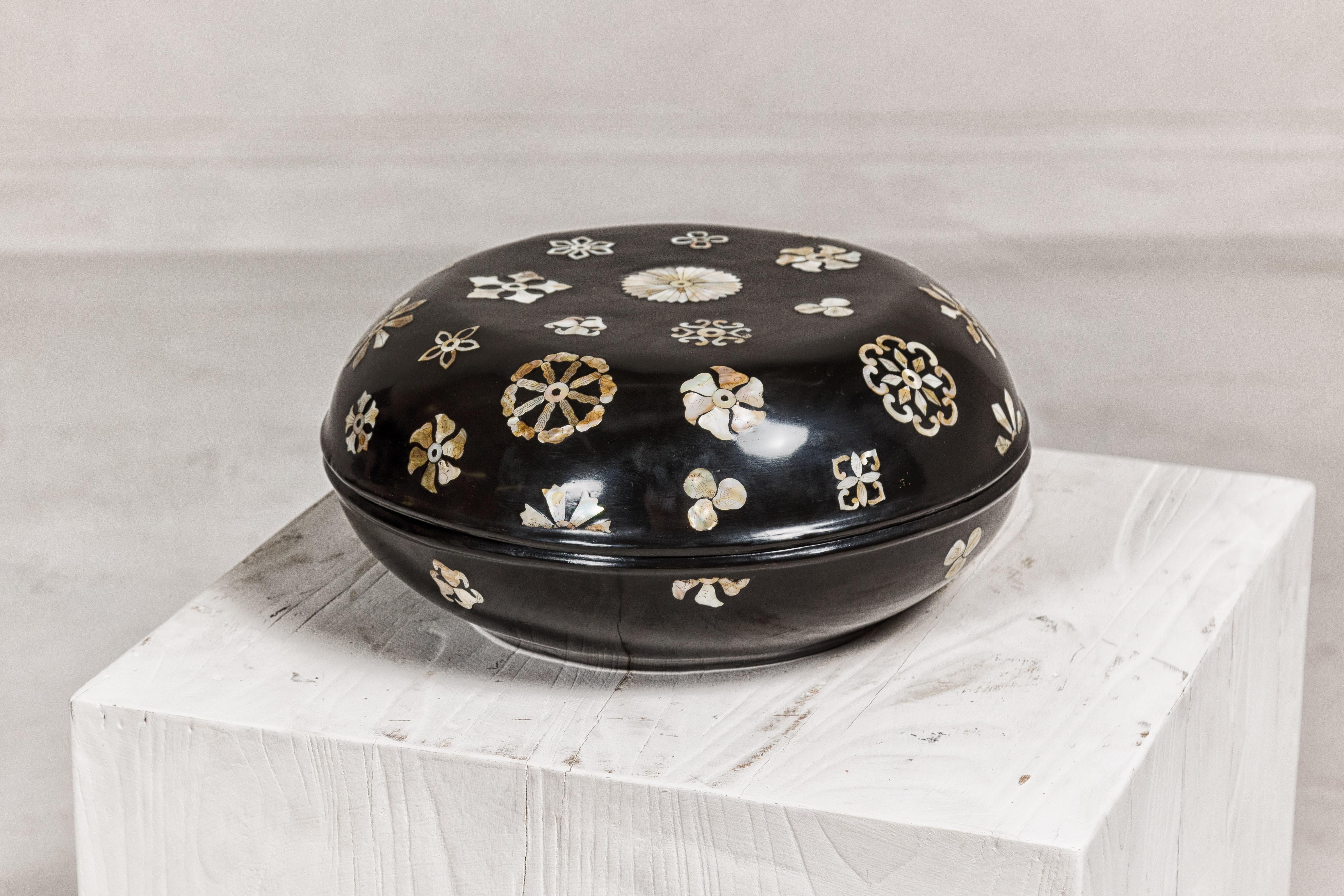 A vintage black lacquered lidded circular box with mother or pearl floral inlaid décor. This vintage Chinese black lacquered box is a testament to traditional craftsmanship, featuring a circular form that is both elegant and functional. The lid is