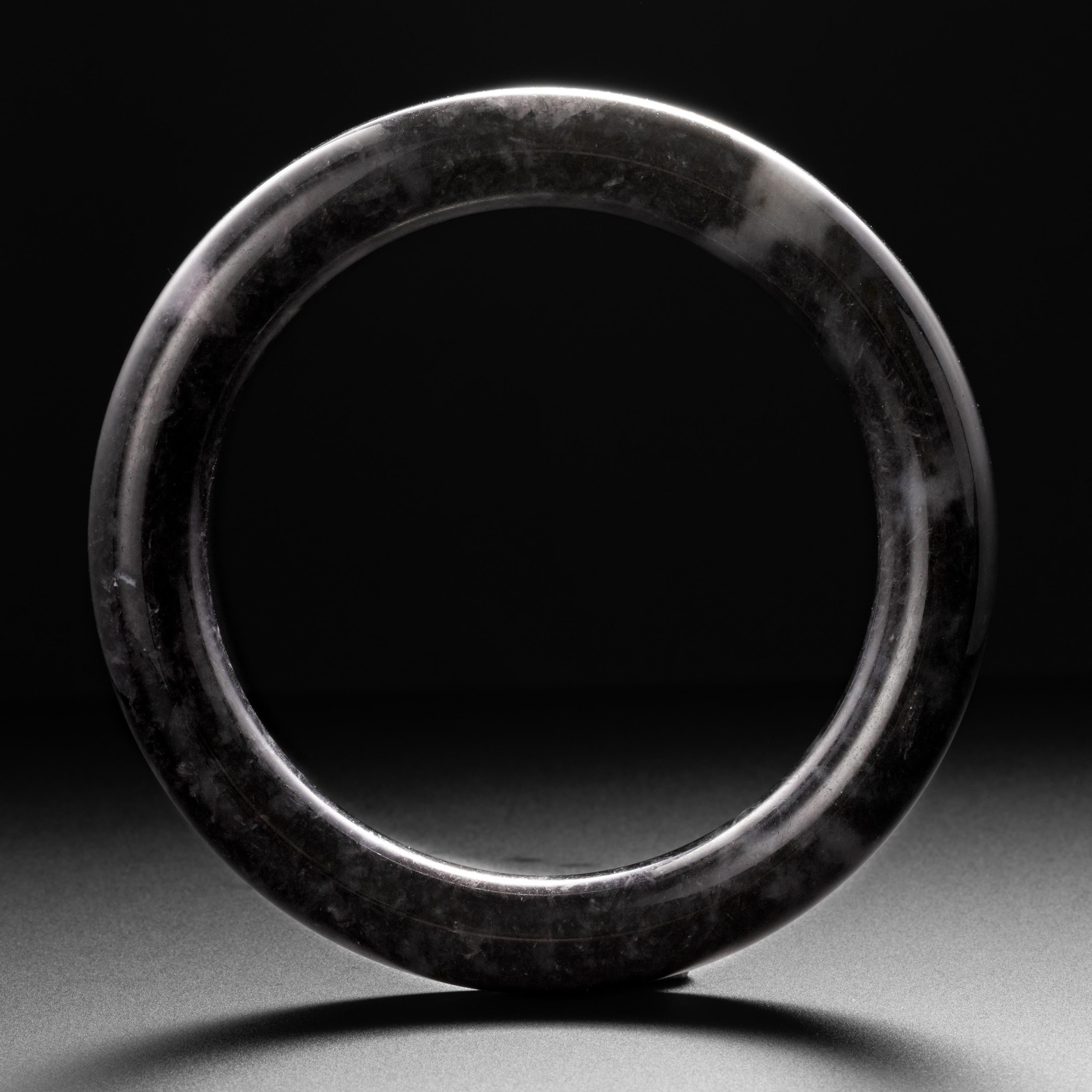 This large jade bangle possesses a brooding beauty. Not quite black, the dark gray jadeite jade bangle is infused in several areas with lighter, more translucent light gray/pale lavender jade. The contrast is striking. The bangle is surprisingly