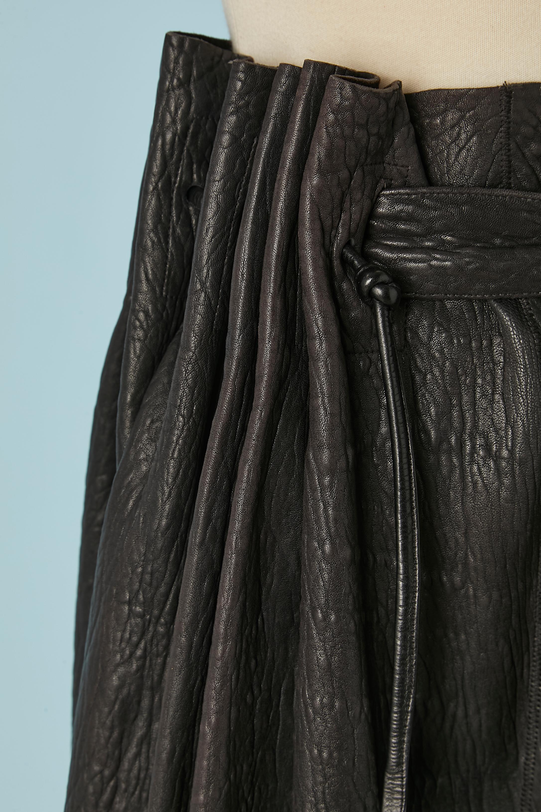 Jupe portefeuille asymétrique en cuir noir  avec sac en cuir sur le devant (attaché à la taille) avec fermeture éclair.  Fermeture par boutons et boutonnière sur le devant. Ceinture en cuir attachée à la jupe et nouée sur le devant. Passant de