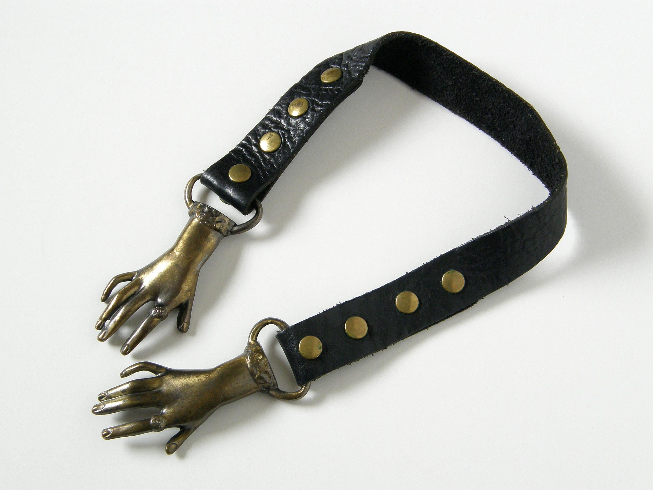 This adjustable, black leather belt has a charming buckle of two overlapping hands that are not quite clasped. They wear rings and bracelets (or perhaps fancy cuff details) and have a romantic, Victorian style with an 
