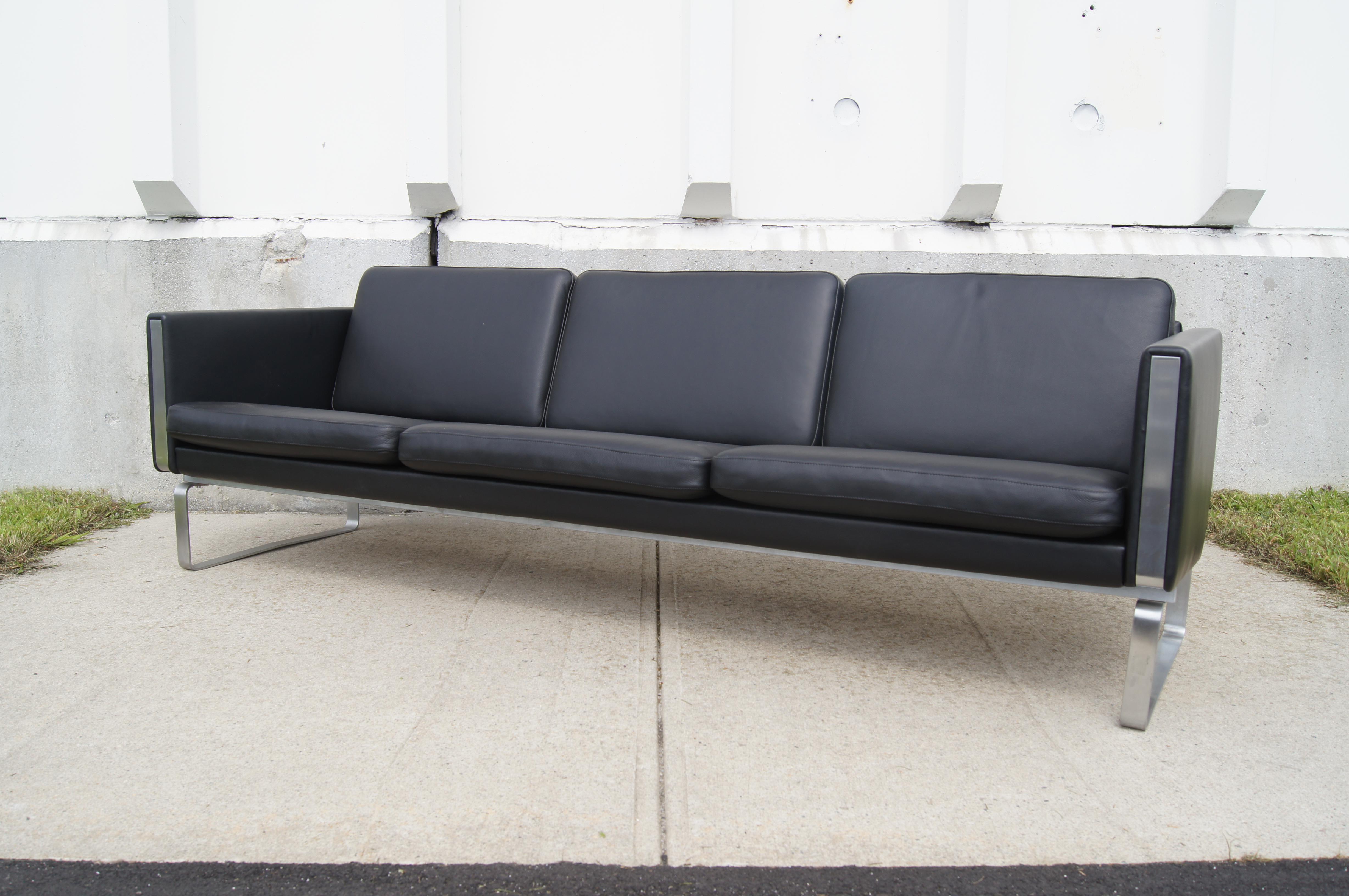 Dieses von Hans Wegner in den frühen 1970er Jahren entworfene, äußerst bequeme Sofa aus schwarzem Leder steht auf einem Sockel aus poliertem Edelstahl, der sich bis zur Vorder- und Rückseite der gepolsterten Armlehnen fortsetzt. 

Dieses zeitlose