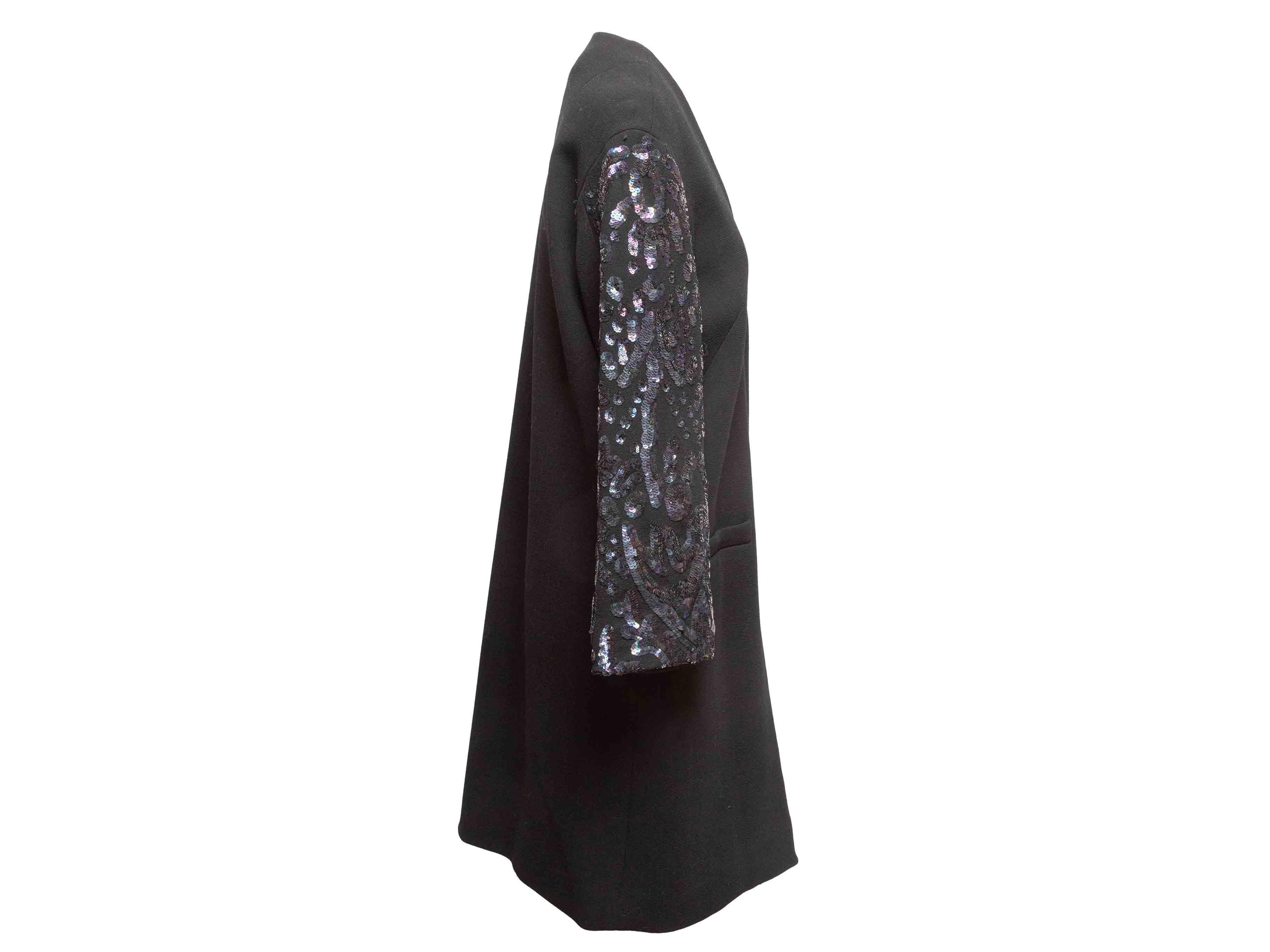 Manteau long sans col noir de Libertine. Col en V. Embellissement des manches par des paillettes. Deux poches sur les hanches. Fermeture par boutons sur le devant. Poitrine 40