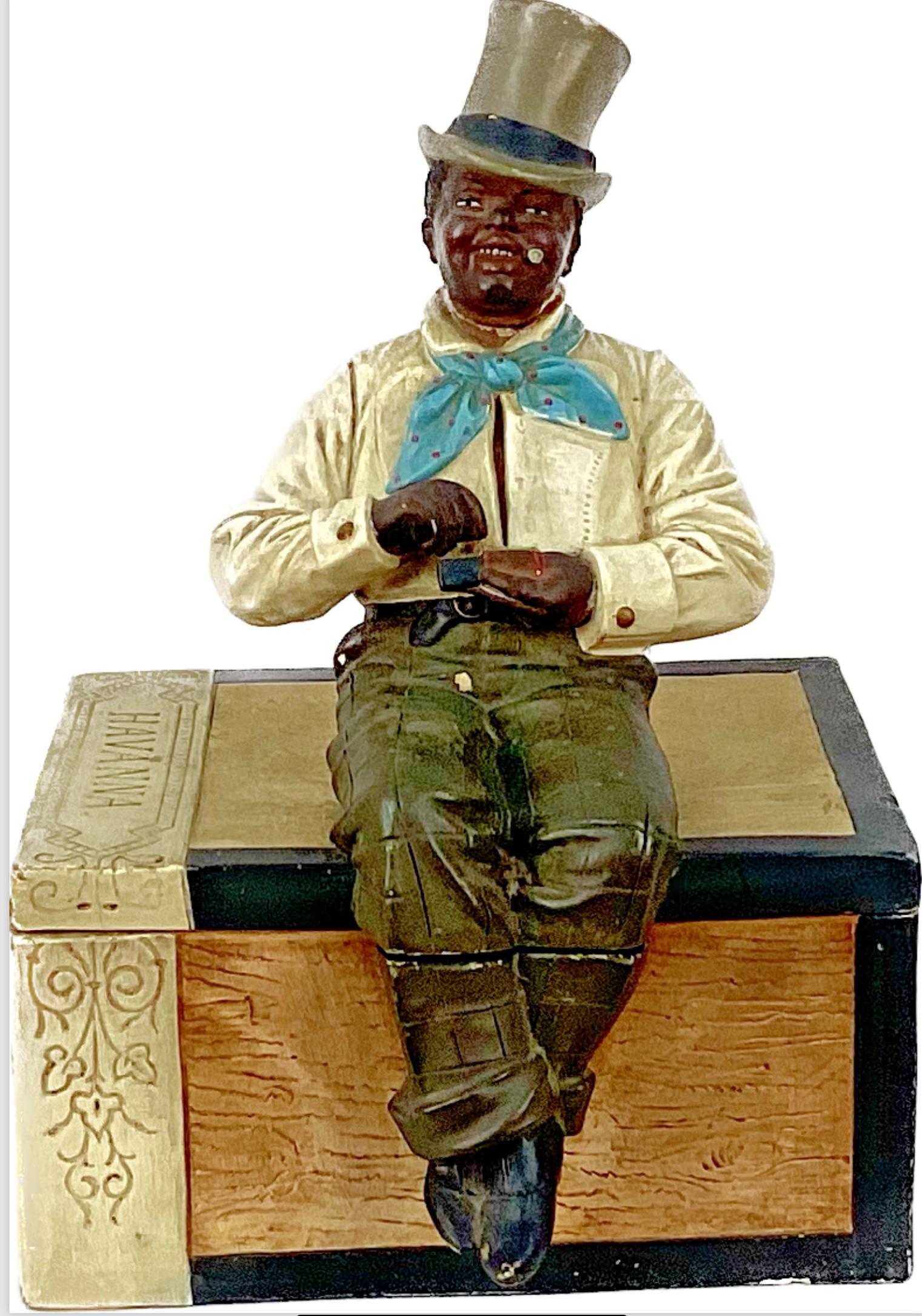 Black Man Ceramic Cigar Jar. Austrian-made, circa 1900, in the form of a wooden cigar box. A nattily dressed black man in top hat sits on the lid with an old-fashioned match box in his hands. Realistic modeling and coloring. This piece is stamped