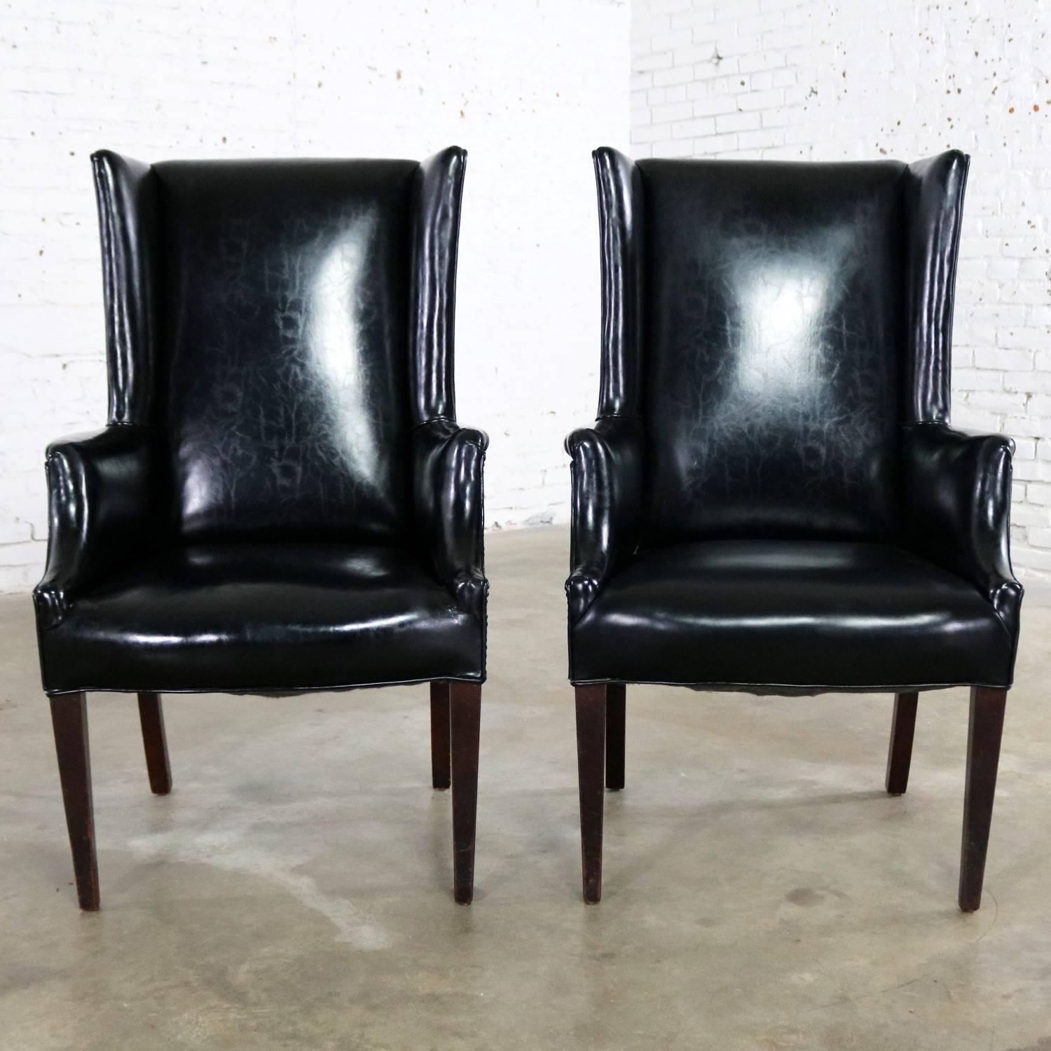 Fabulous pair of black faux leather or Naugahyde wingback chairs with nailhead detail in an Art Deco or Hollywood Regency styling. This pair is in wonderful vintage condition, sturdy with age appropriate wear. The upholstery on one chair is without