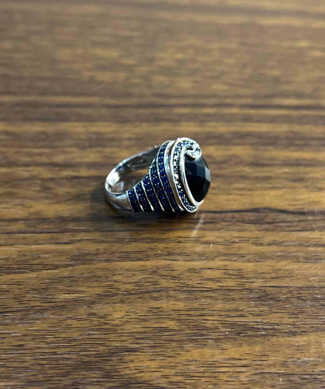Elegant Black Onyx and Sapphire White Gold Ring. This striking ring features a large faceted black onyx stone. The onyx is surrounded by a halo of small blue sapphires, adding a touch of color and elegance. The band is intricately designed with a