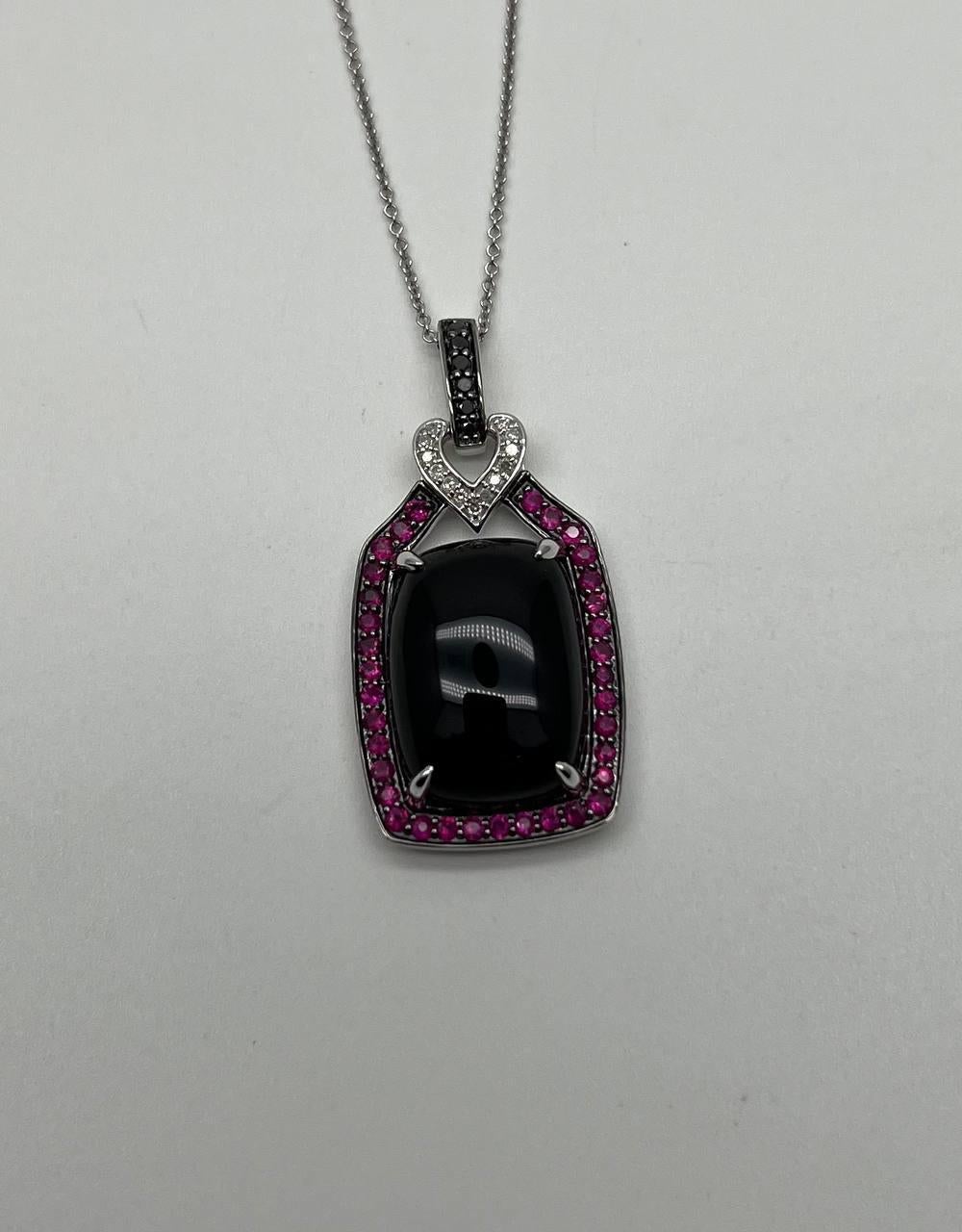 Pendant necklace featuring a central black cabochon Onyx framed by a border of round pink red rubies. The pendant is suspended from a chain with a heart-shaped design adorned with small white diamonds.
14K White Gold
0.15 CTW Diamonds
Black Onyx and
