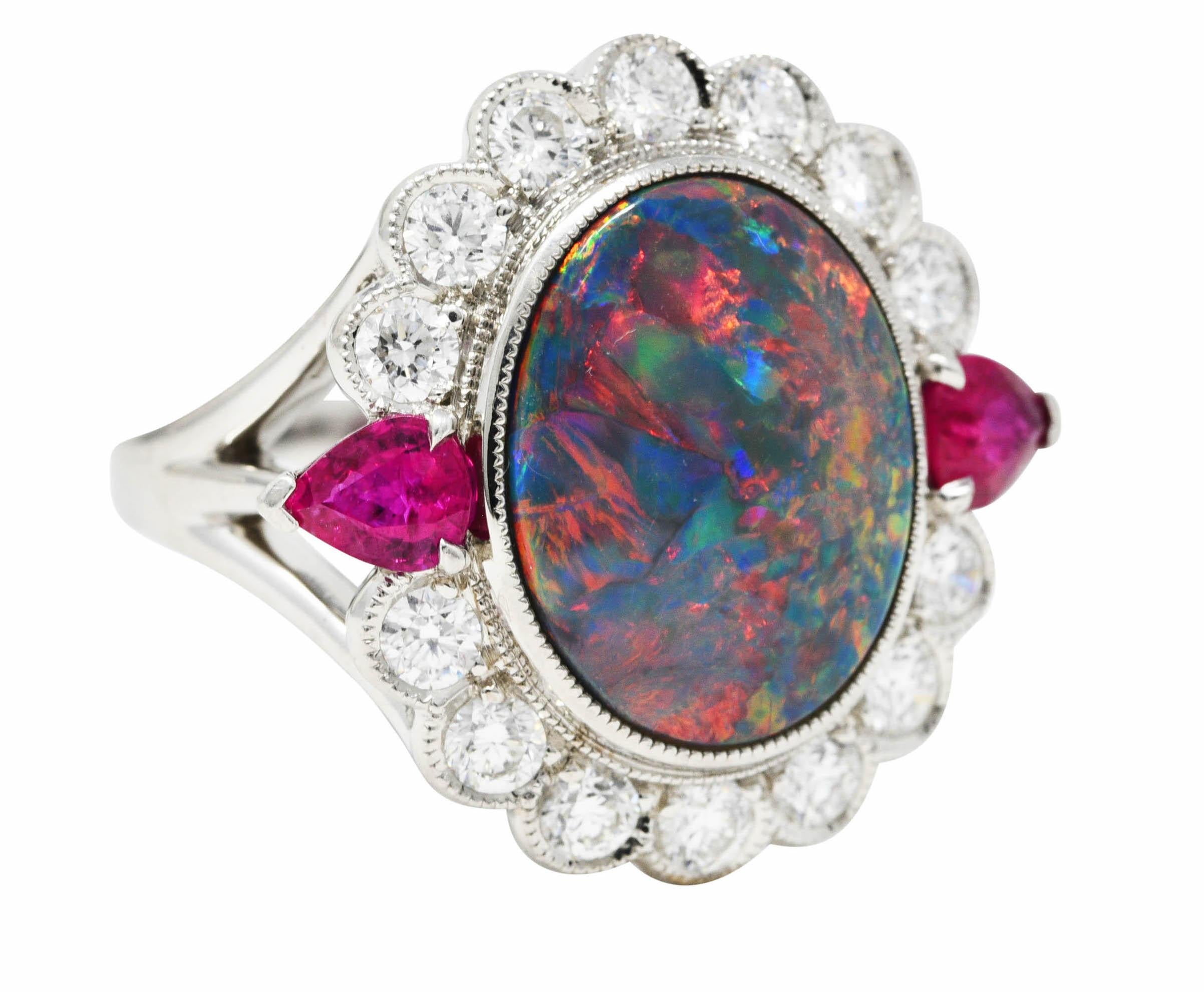 Statement cluster features on oval black opal cabochon measuring approximately 15.0 x 11.8 mm. Opaque dark body color and very strong spectral play-of-color with broad flashes intermittent by pinfire. Bezel set and flanked by two pear cut rubies