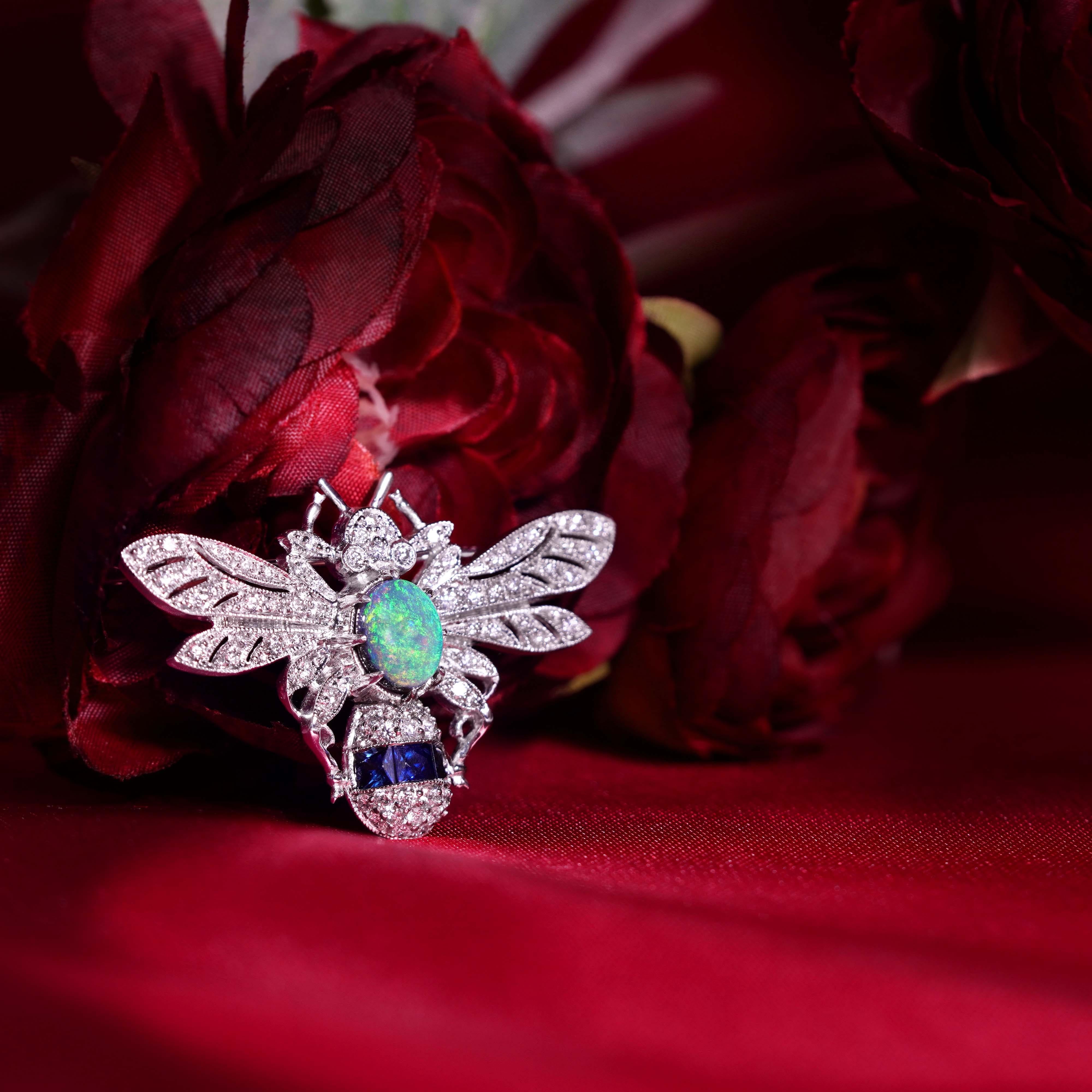The Black Opal, Sapphire, and Diamond Art Deco Style Bee Brooch features a mesmerizing black opal as the body, showcasing its mysterious, iridescent hues. The wings, adorned with sparkling diamonds, add a touch of brilliance, while sapphires