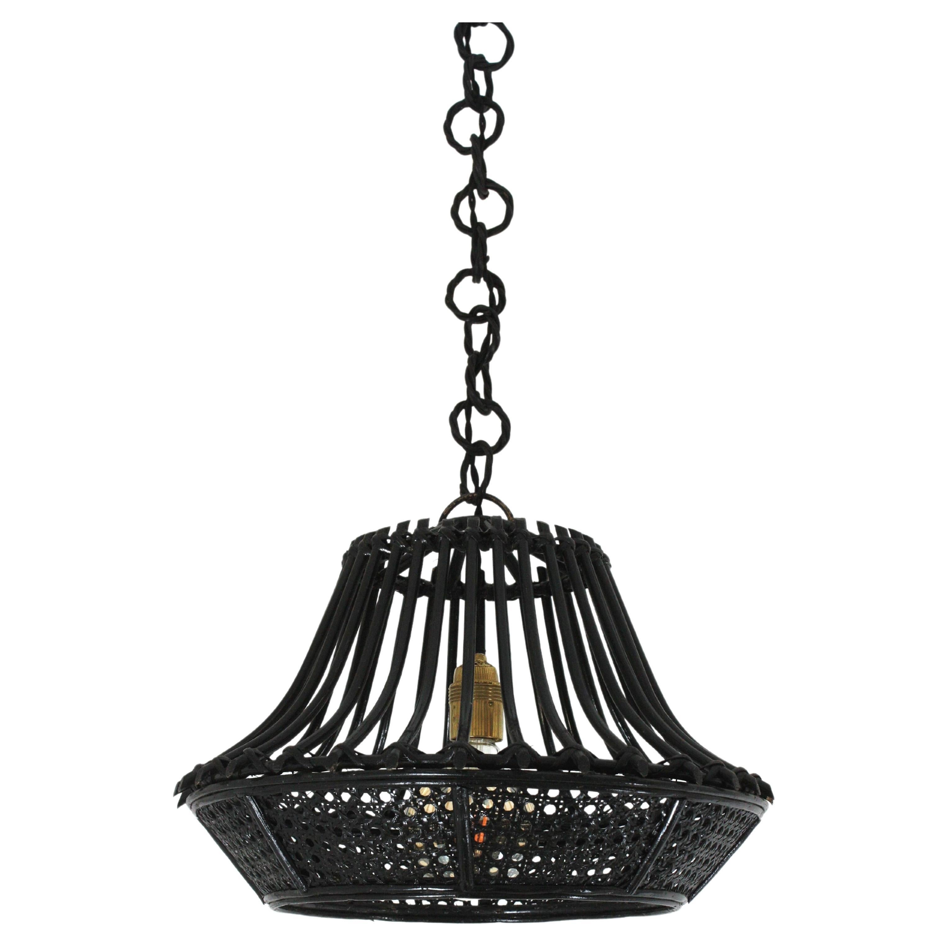 Pendente / lampadario a forma di pagoda in rattan e filo di vimini patinato in nero, Francia, anni '60.
Questa lampada a sospensione di metà secolo presenta un paralume conico in vimini con canne verticali in rattan che pendono da una catena fatta
