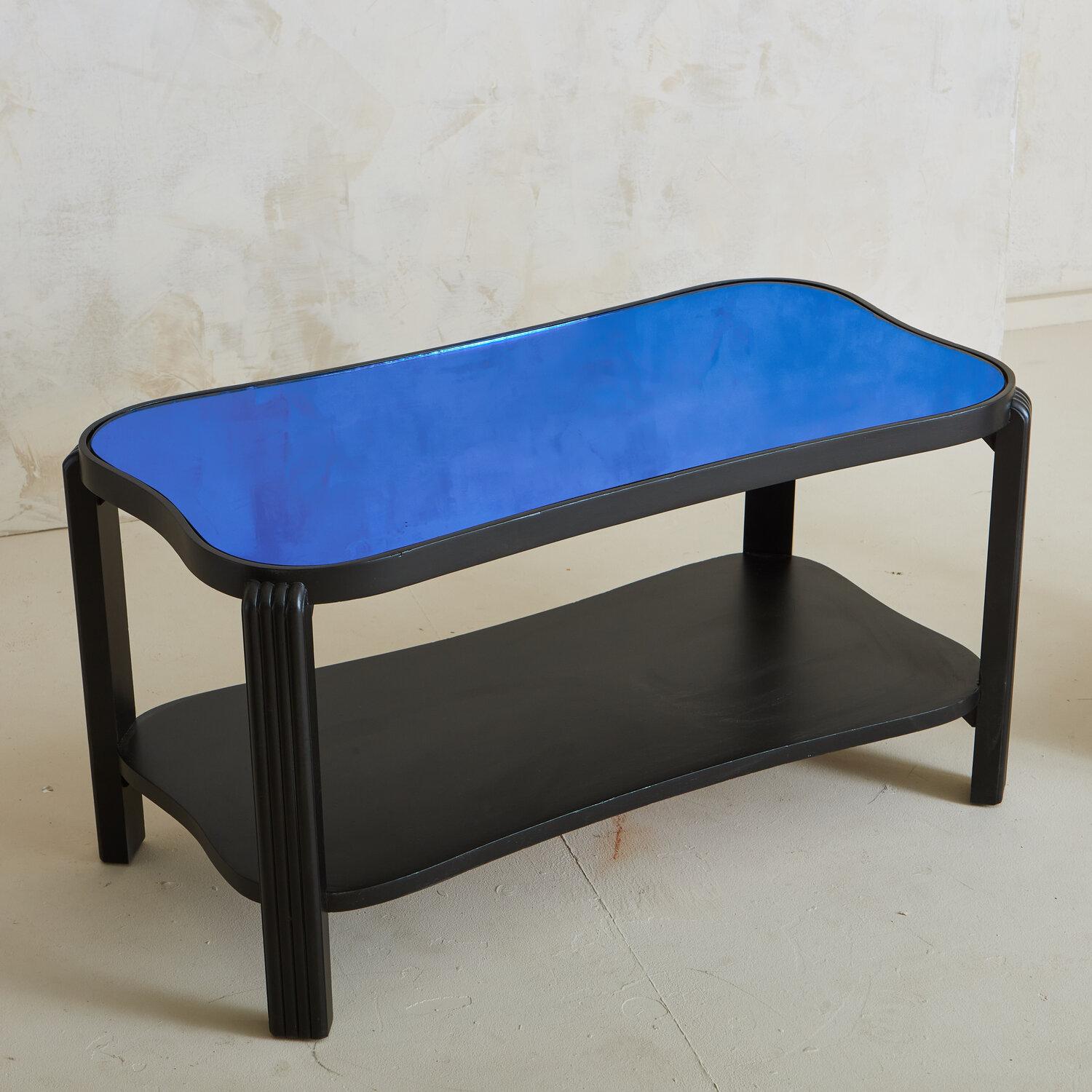 A black painted wood coffee table with rounded edges and fluted legs. This table features a vibrant blue reflective glass tabletop and two-tiers perfect for styling books and trinkets. It has one matching side table.