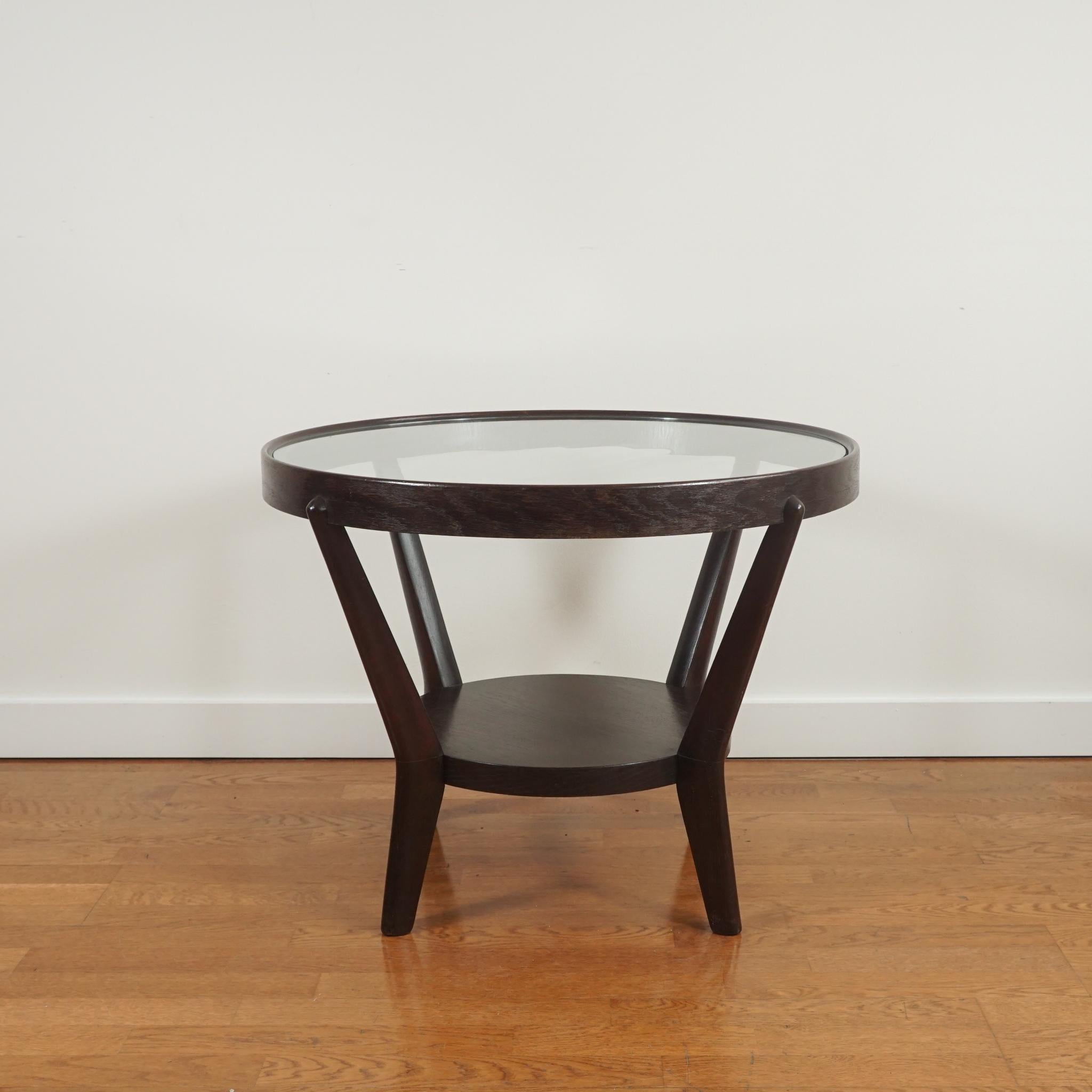 The round glass top wood side table, shown here, defies its age. Made in the 1950s, it has been gently restored to enhance the original black patinated finish. The glass top with bottom shelf makes the table appear lighter and perfectly suited for