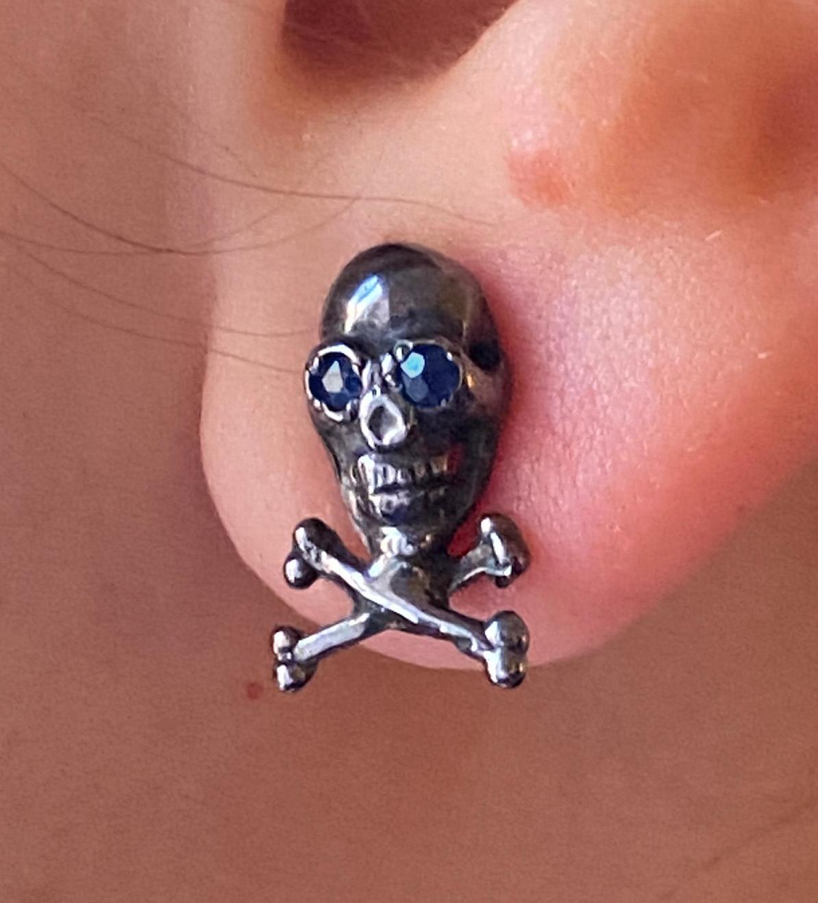 Single men's stud earring in black rhodium-plated sterling silver, designed in the shape of a skull with crossed bones beneath it. The skull features sapphire blue eyes and represents a skeleton, evoking a strong and rebellious spirit with its bold,