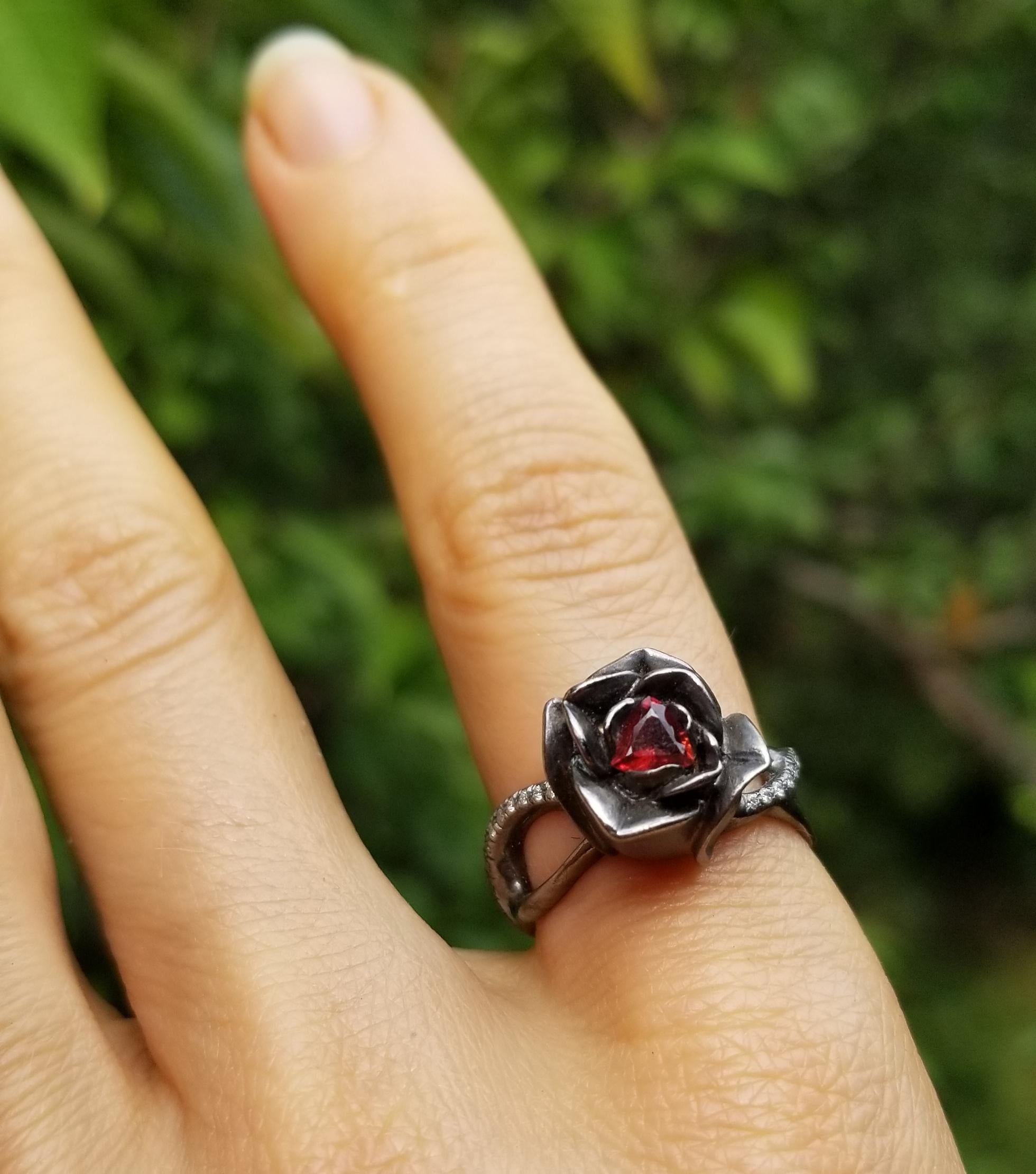 A symbol of rebirth and hope, this stunning black rose ring was hand-carved in wax and cast in Sterling silver, then set with a perfect blood-red trillion garnet in the center.
White diamonds frame the flower on either side and cascade down the