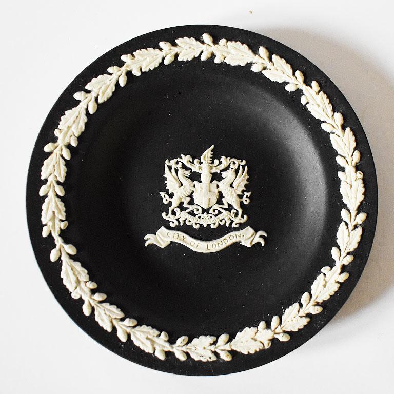 Round black jasperware dish by Wedgwood. A lovely piece with white decorative accents atop a black background. At the center, it features a crest with the City of London at the bottom. This would make for a great accent piece on a nightstand as a