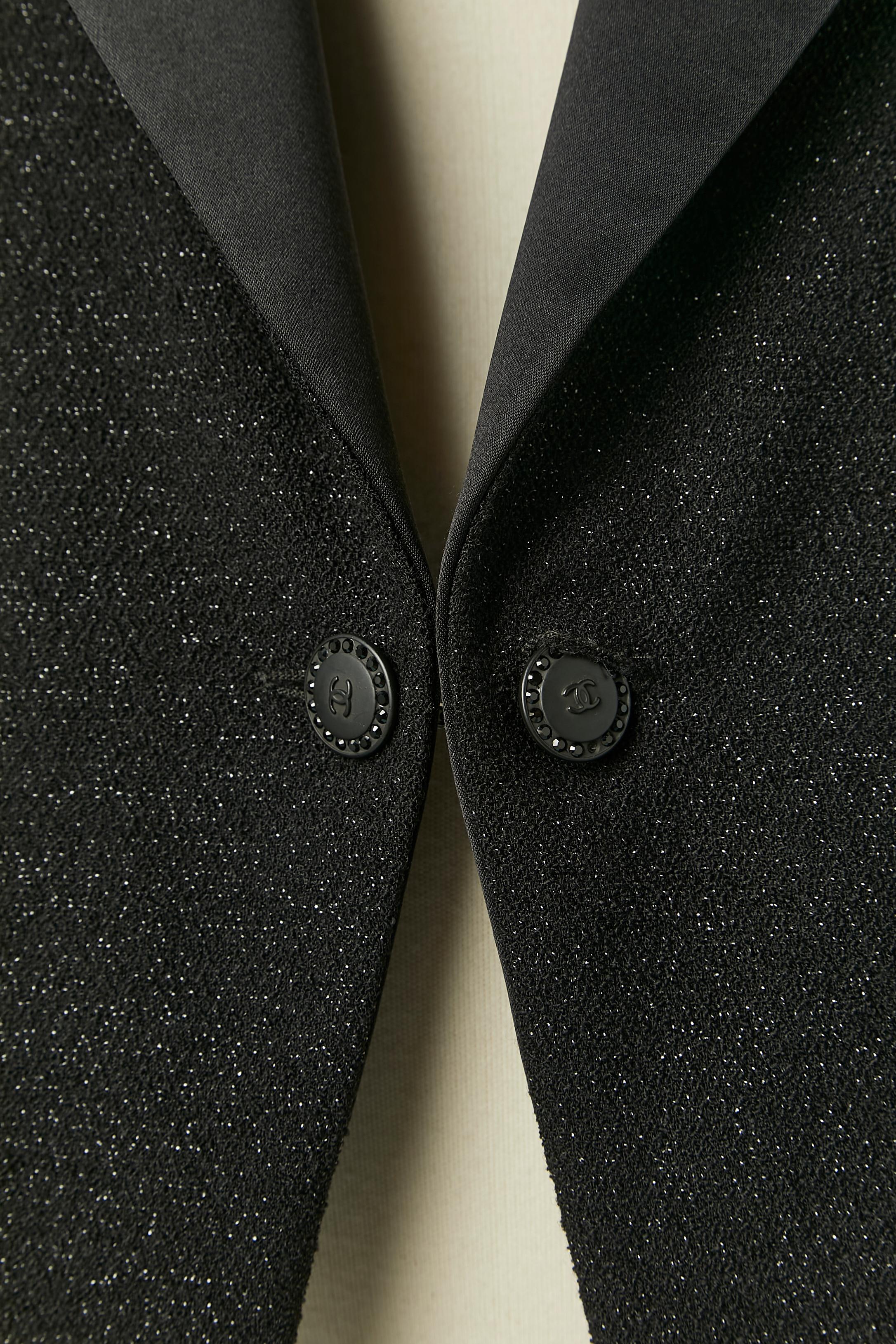 Chaqueta de lana negra brillante de borde a borde con cuello de seda. Composición del tejido principal: 85% lana, 15% poliamida. Cuello de seda. Forro de marca: 96% seda, 4% elastán. 
Hombreras. Cierre con 2 botones de marca en la parte delantera