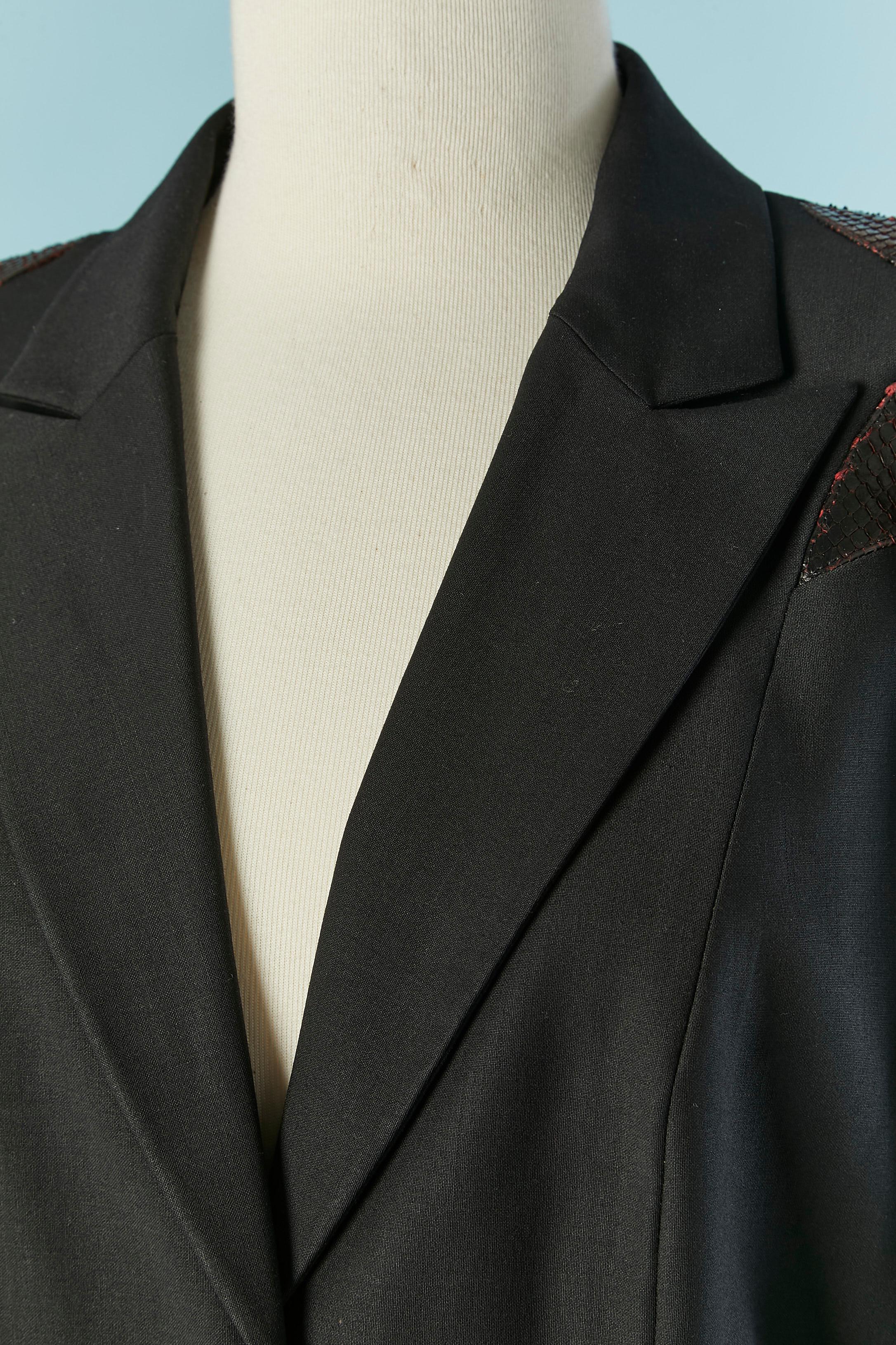 Giacca monopetto nera con applicazione sulla spalla. Composizione del tessuto principale: 60% lana, 30% rayon, 6% seta, 4% lycra. Chiusura a scatto nascosta sulla parte centrale del davanti. Fodera in seta rossa. Tasca su entrambi i lati. Spacco sui