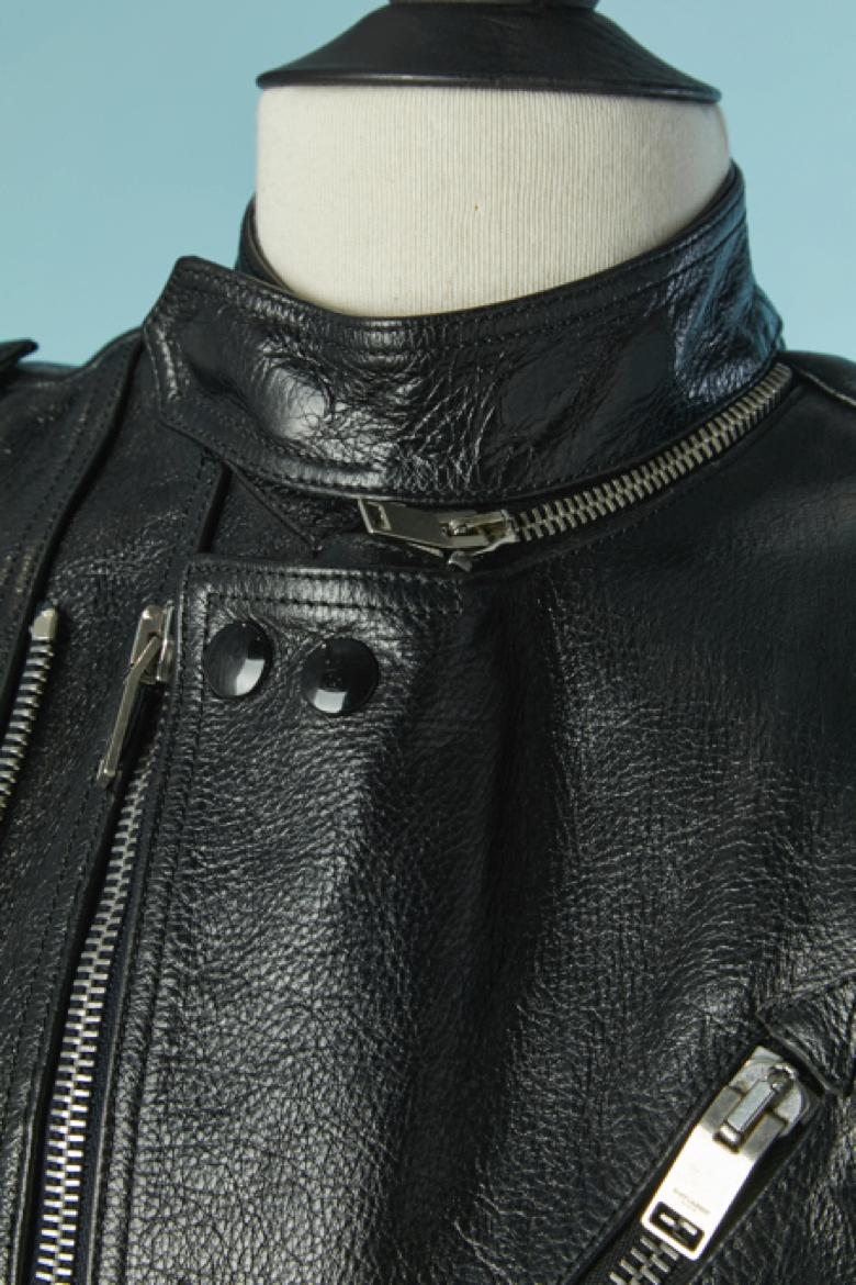 Schwarze ärmellose Bikerjacke mit leichter Polsterung. Reißverschluss in der Mitte der Vorderseite + Druckknopf an der Oberseite. Einige Teile sind abgesteppt. Gürtel mit Schnalle. 3 Außentaschen und 2 Innentaschen. Schnürsenkel auf dem