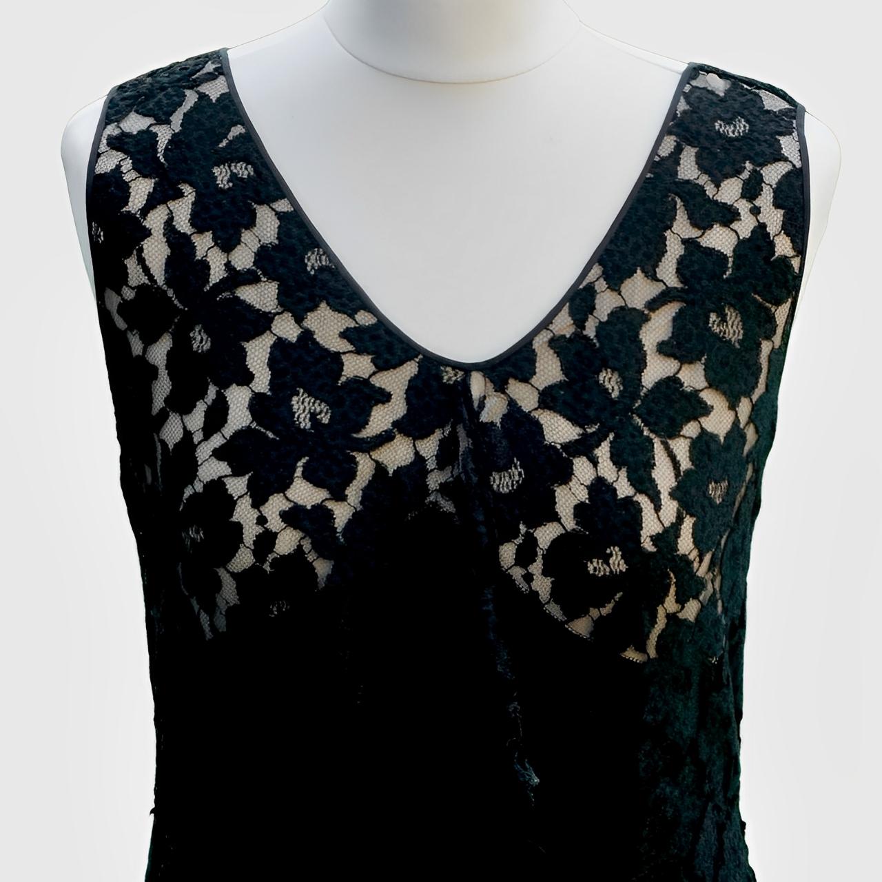 Magnifique robe en dentelle noire sans manches au motif fleuri, avec un volant sur le devant. La robe s'évase dans le bas.

Le corsage est doublé dans une matière nude, la robe principale est doublée en noir. La fermeture latérale est munie d'un