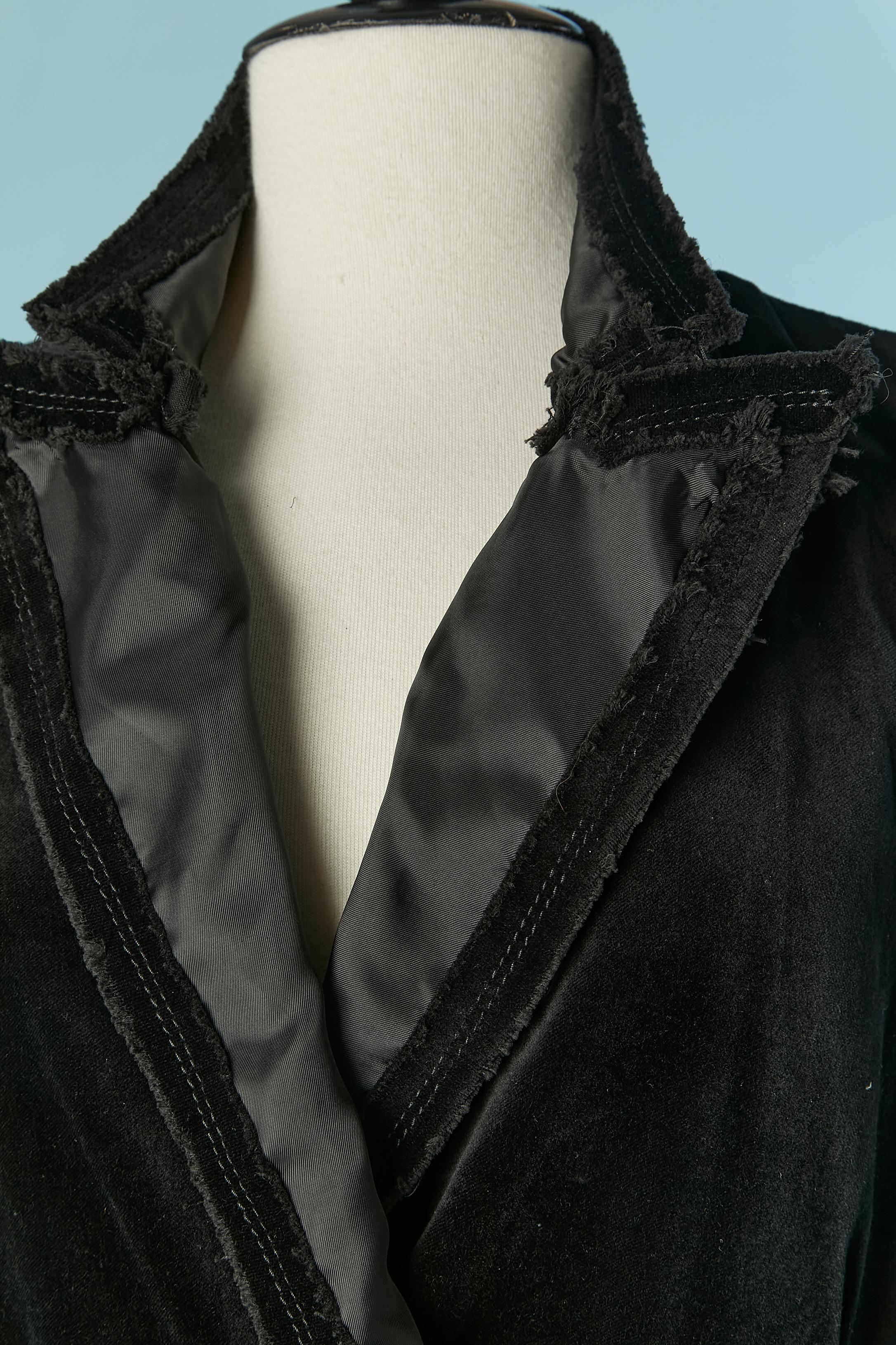 Schwarzer ärmelloser Samtmantel mit rauer Schnittkante. Zusammensetzung Samt: 100% Baumwolle. Zusammensetzung des Futters (nur auf der Vorderseite des Mantels): 100% Polyester. Paspelierung auf der Innenseite des Rückens. 
Druckknopfverschluss auf