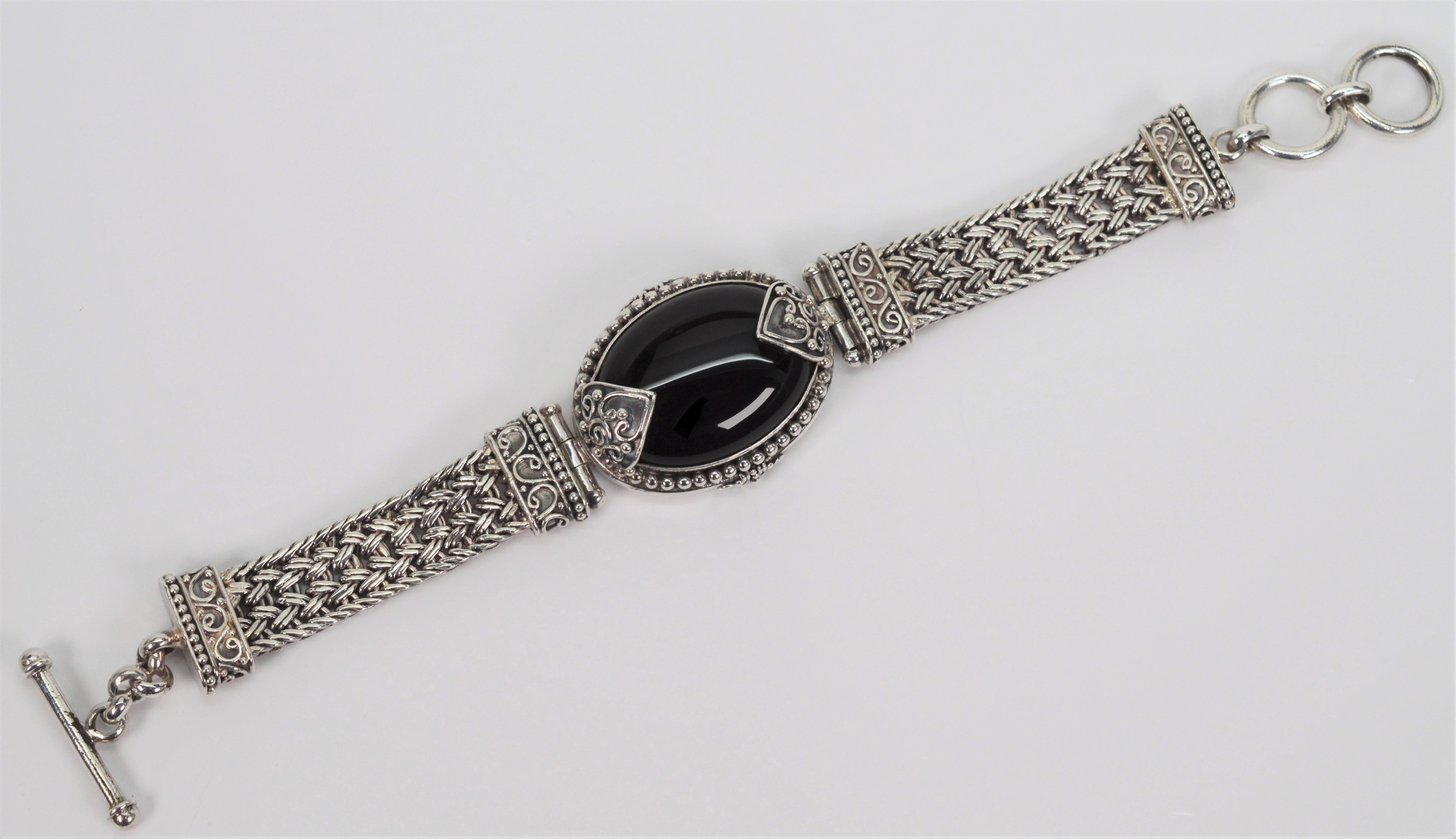 A woven sterling silver band with outstanding artful details in silver wire filigree and silver drop chain findings surround the a 22 x 34 mm oval black stone cabochon featured on this vintage style statement bracelet. Seven inches in length with a